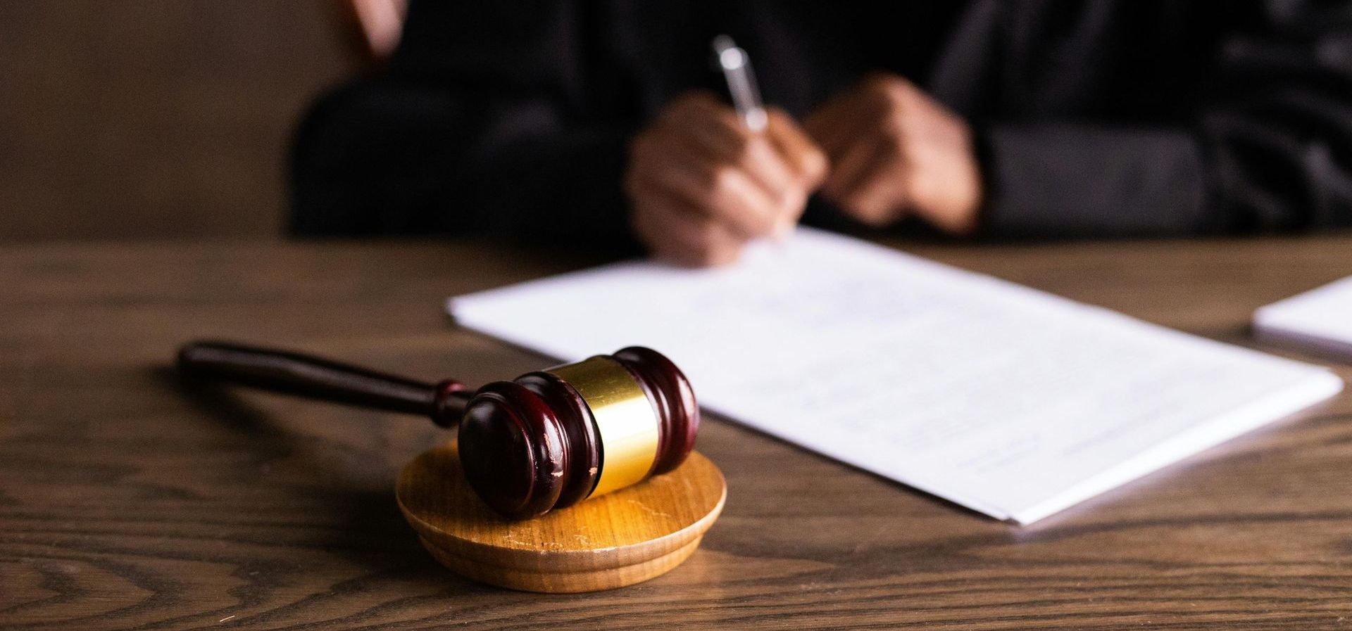 A judge 's gavel is sitting on a wooden table next to a person writing on a piece of paper.