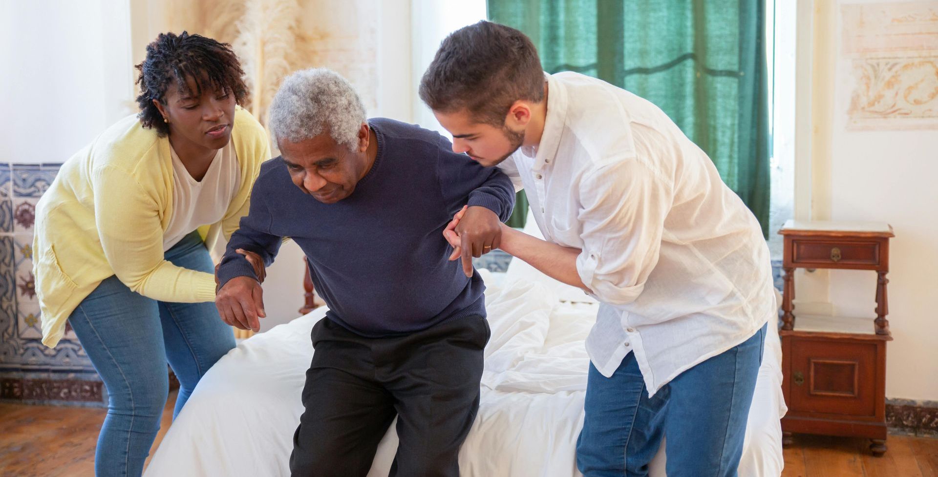 A man and two women are helping an elderly man get out of bed.