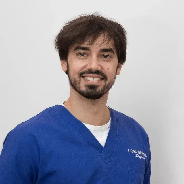 Un professionista in camice blu con maglietta bianca sorride su uno sfondo bianco uniforme.