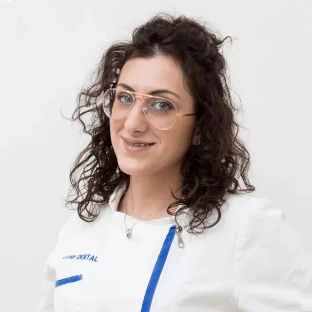 Un professionista in camice bianco con bordi blu, sorridente, con capelli castani ondulati e occhiali dalla montatura chiara.