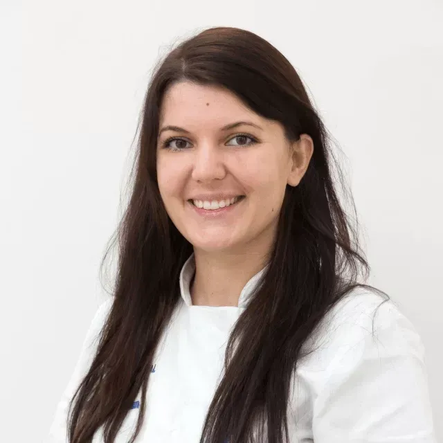 Una persona con lunghi capelli scuri, sorridente, che indossa un'uniforme bianca su uno sfondo bianco uniforme.