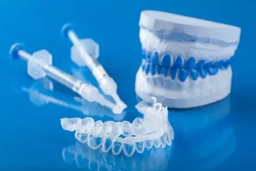 Modello di impronta dentale, vassoio per allineatori in plastica trasparente e due siringhe di gel sbiancante su sfondo blu.