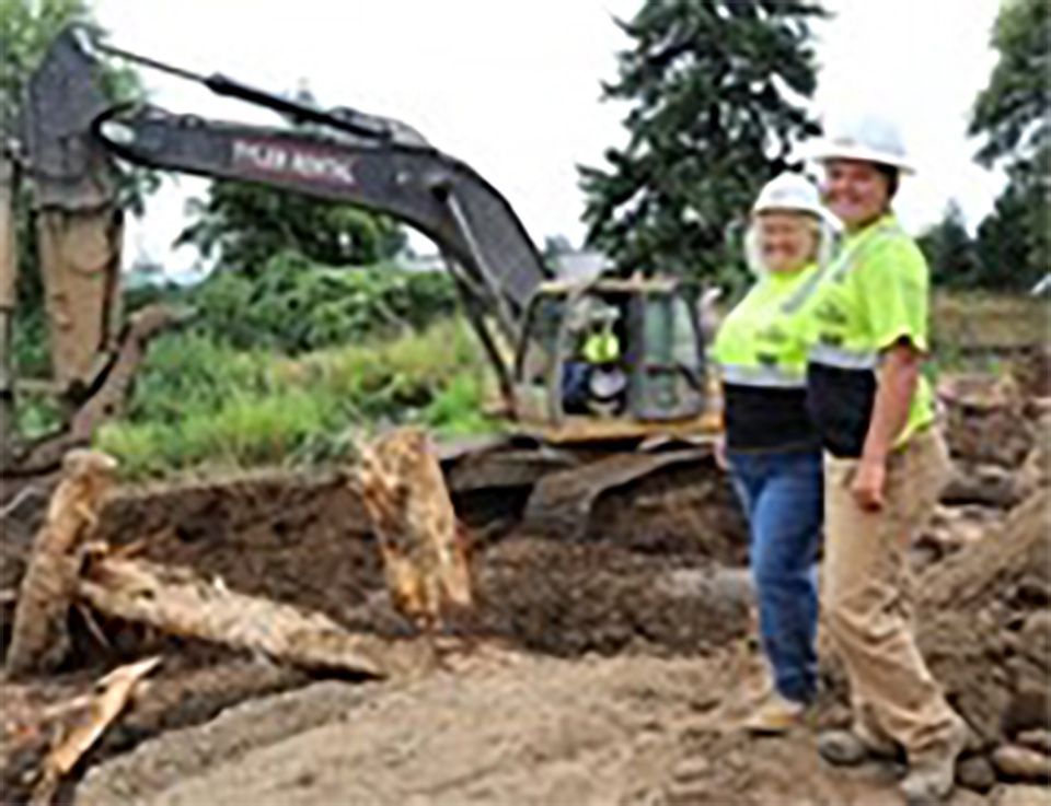 Two women are standing in front of a large excavator.