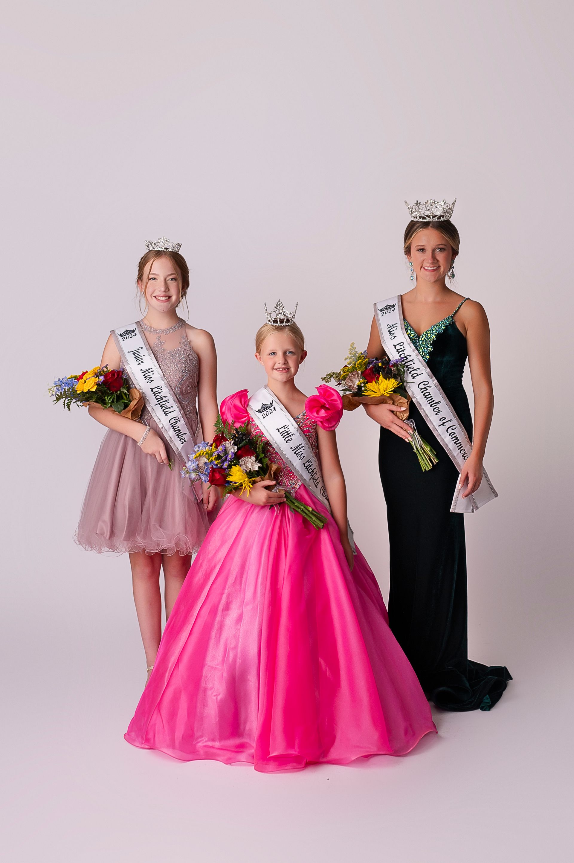 Three young girls are standing next to each other holding flowers and sashes.