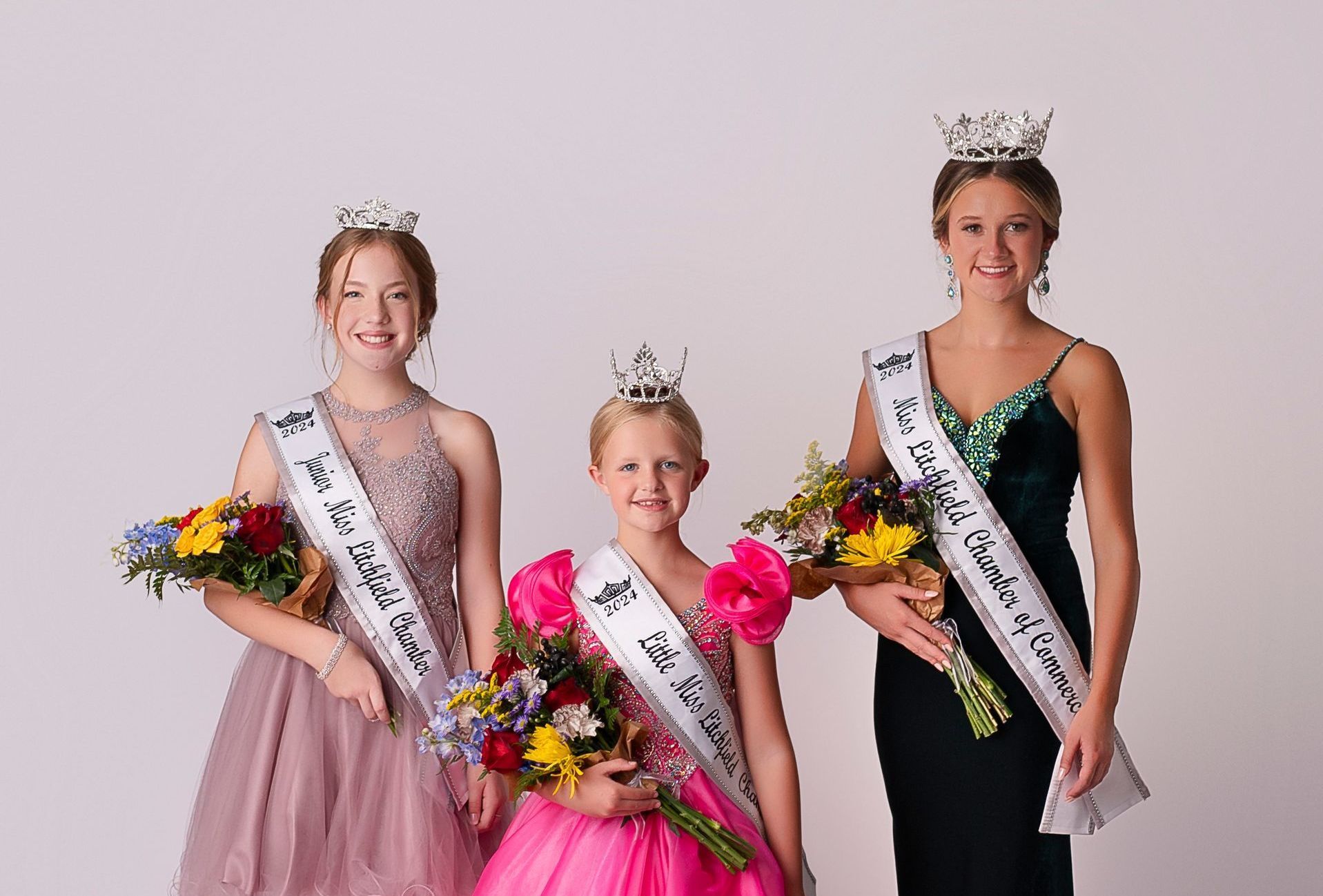 Three young girls are standing next to each other holding flowers and sashes.