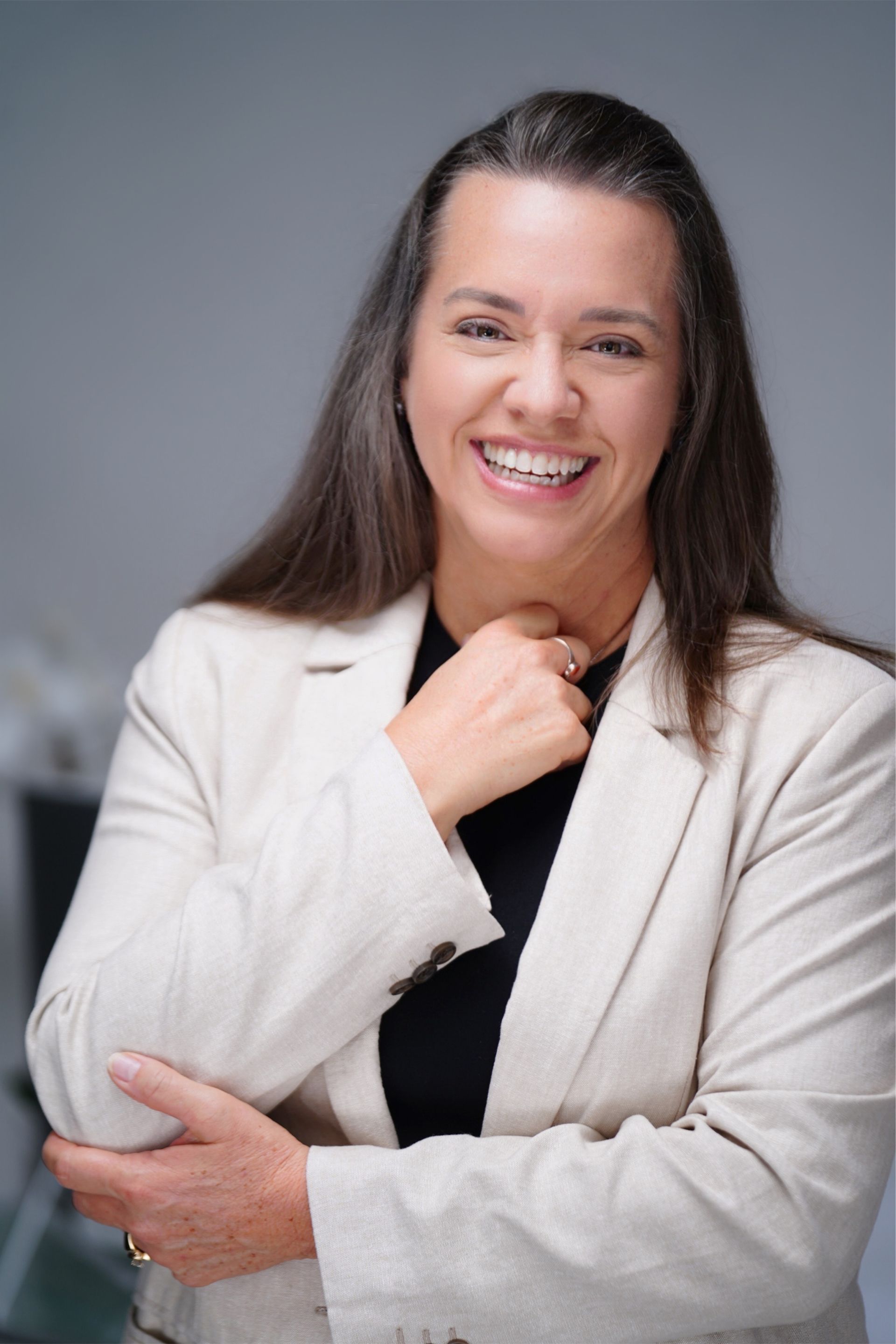 Woman in blazer smiling, hand near chin, with long brown hair.