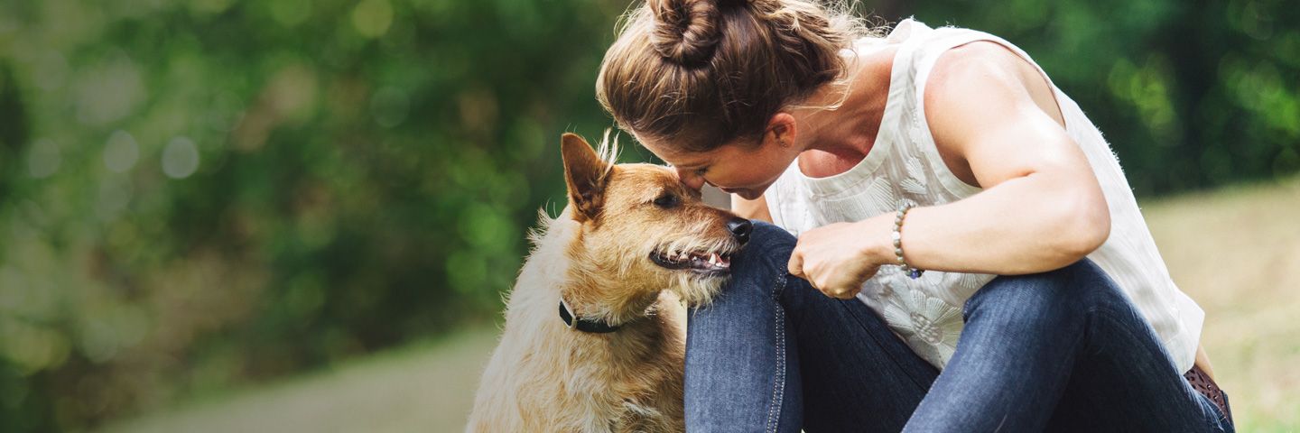 A woman is kneeling down next to a dog and kissing it on the nose.