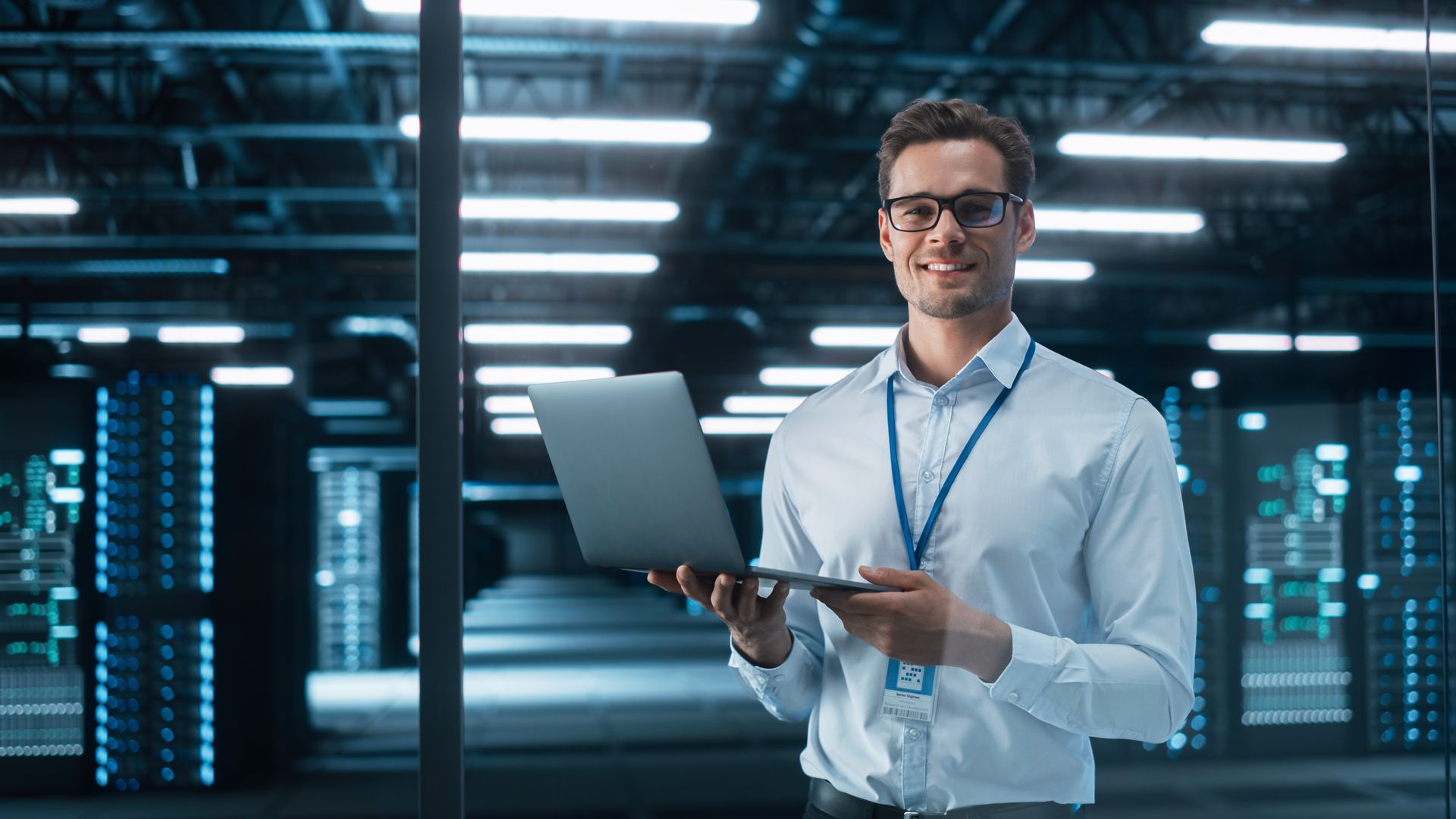 Smiling network administrator standing inside a data center and holding a laptop