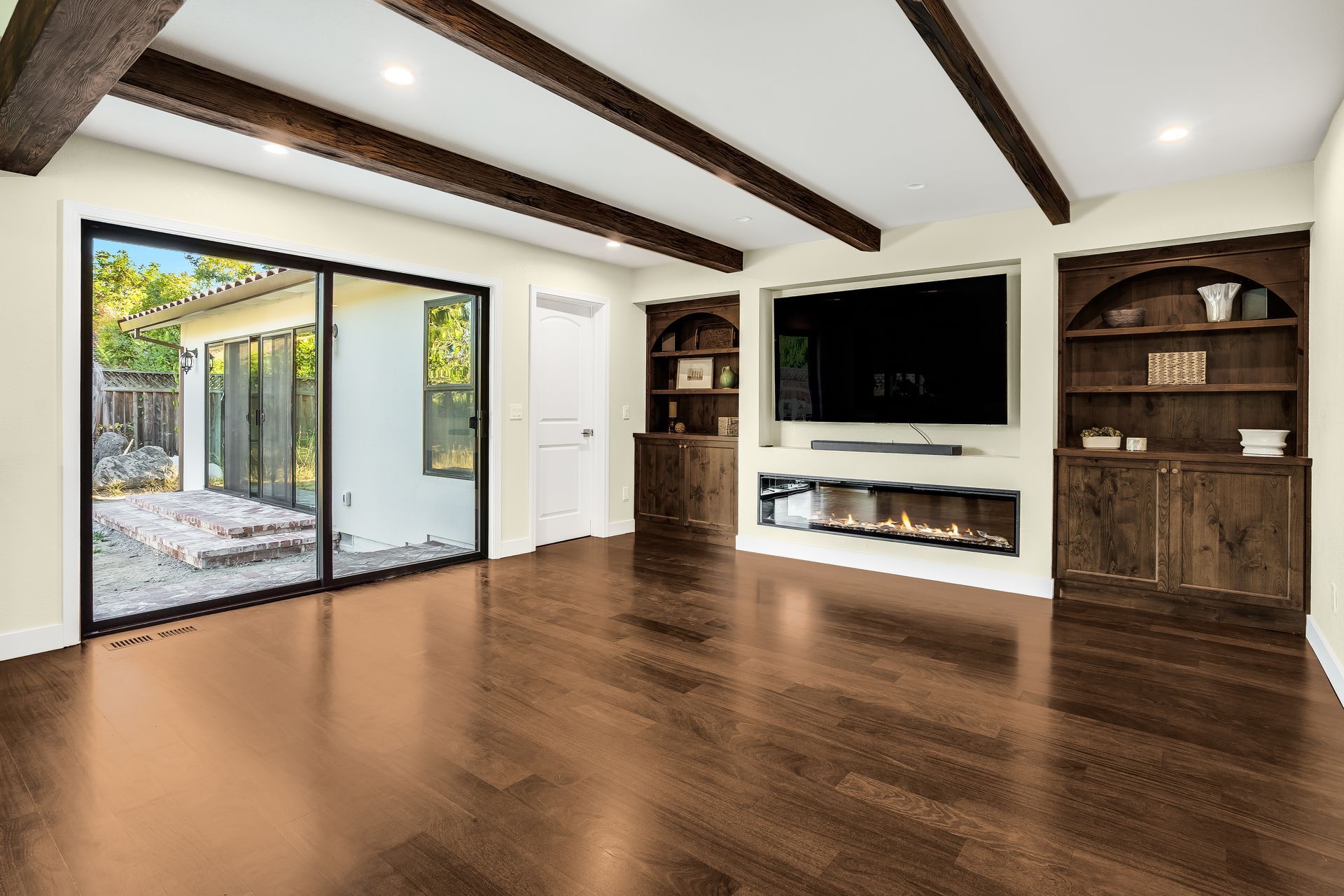 Empty living room with dark wood floors, large TV, fireplace, built-in shelving, and sliding glass doors to the backyard.