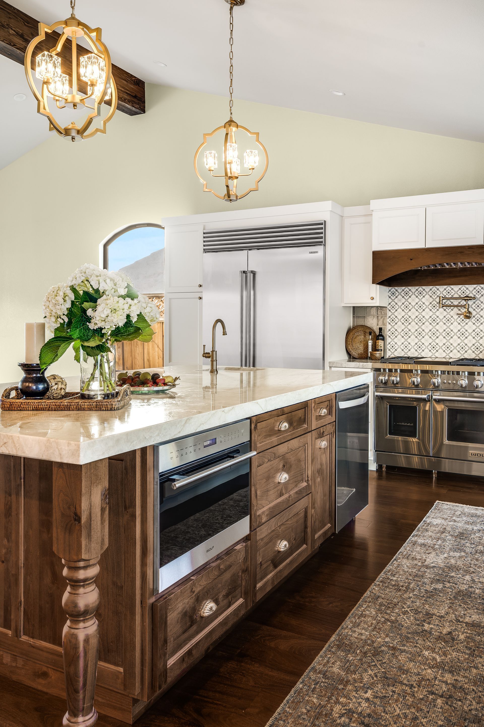 Upscale kitchen with wooden island, built-in appliances, marble countertop, gold light fixtures, and stainless steel oven.