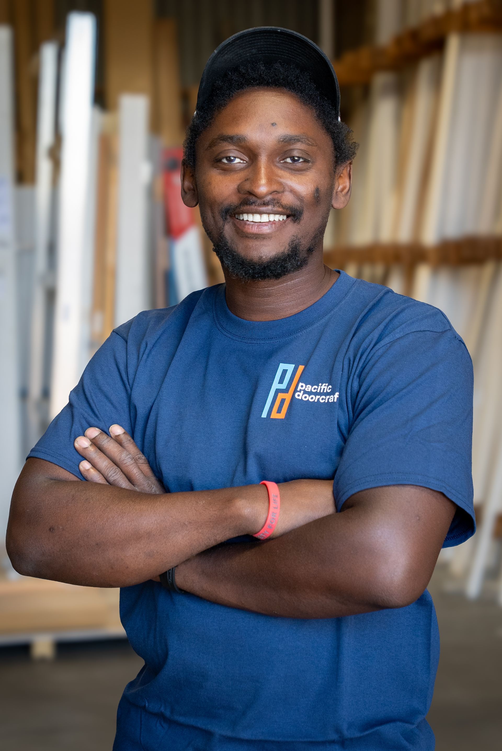 Man with crossed arms smiling in a workshop, wearing a blue shirt and baseball cap.