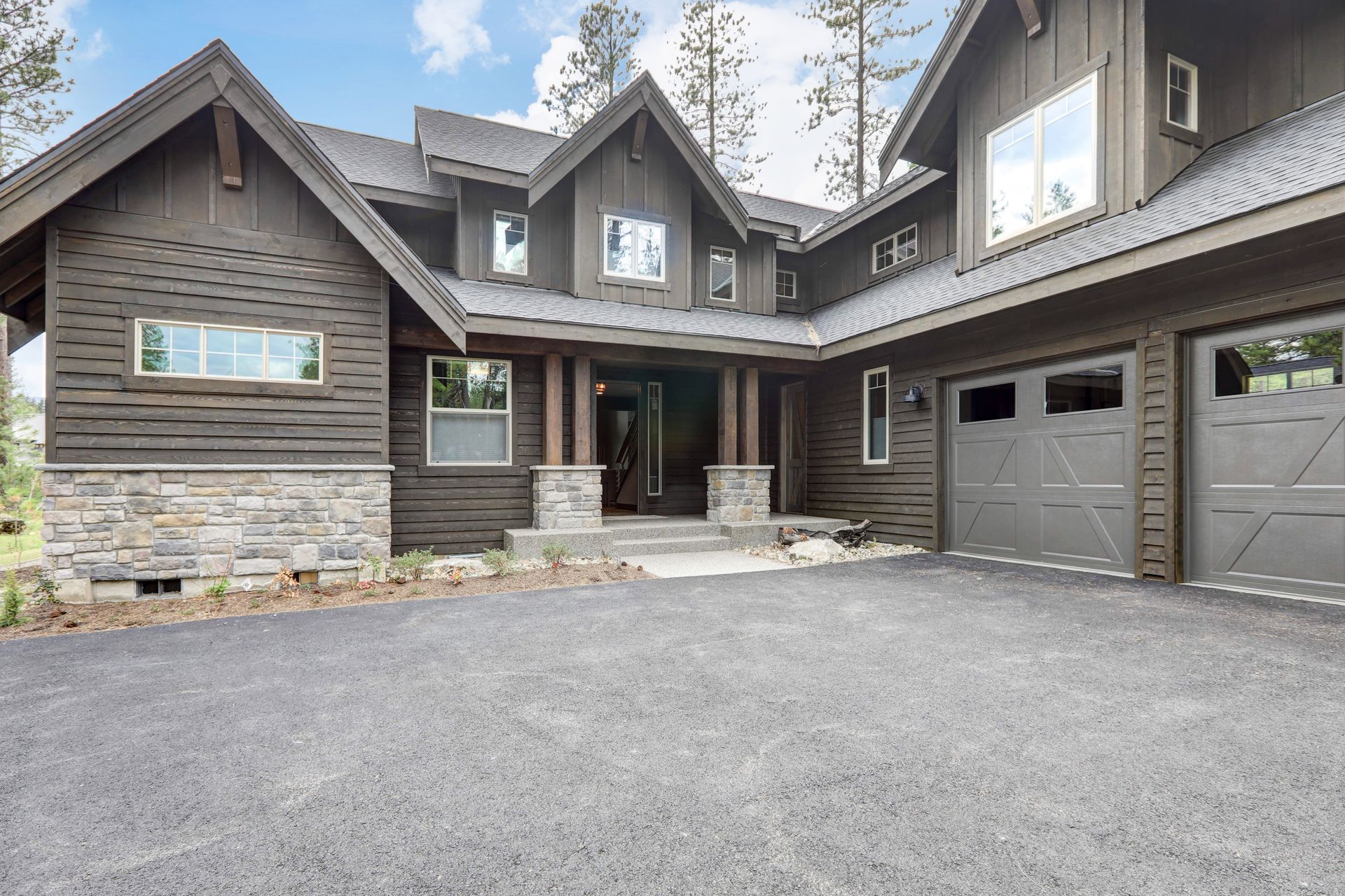 Brown wood-sided house with stone accents, gray garage doors, and gravel driveway under a cloudy sky.