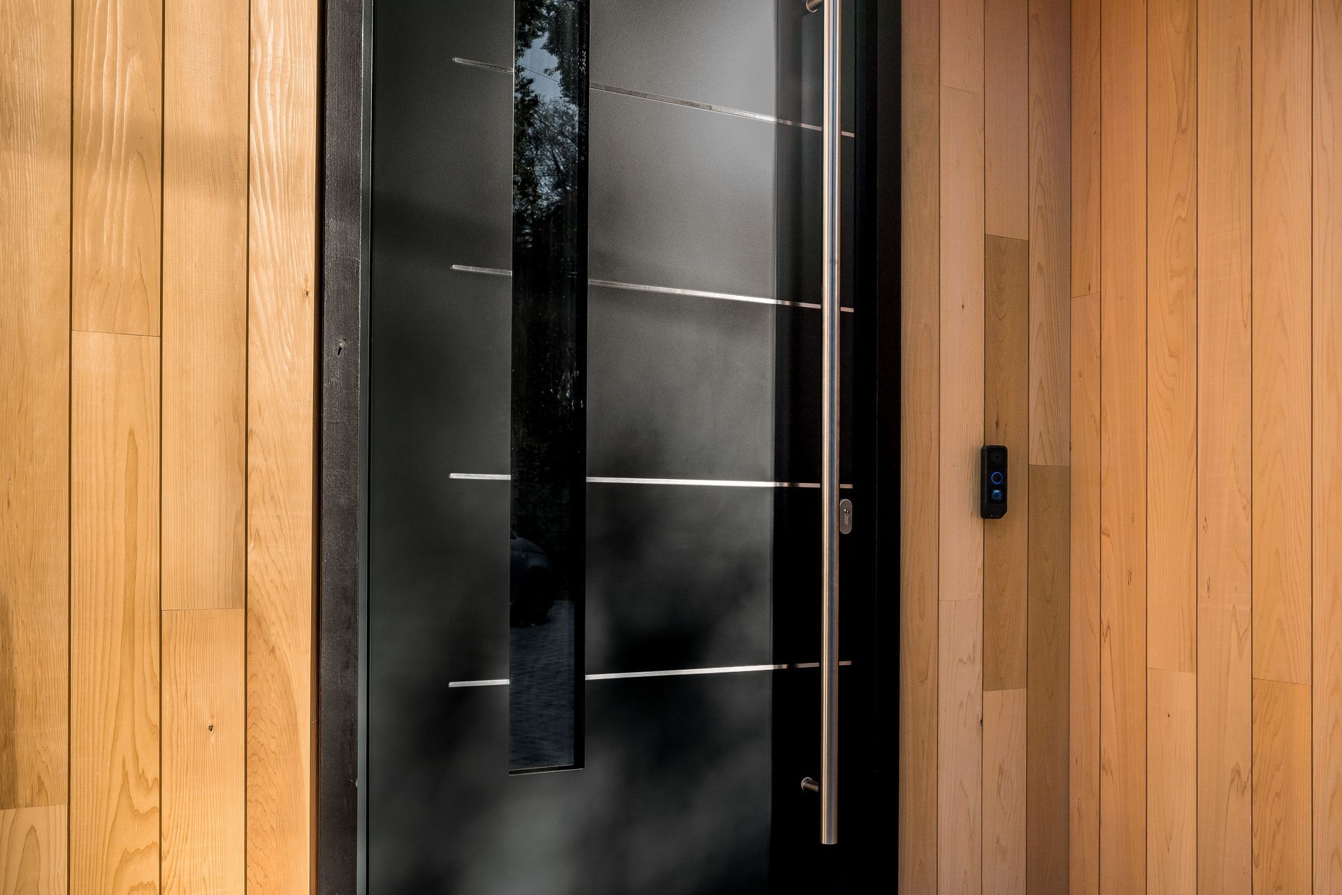 A black door with a stainless steel handle is on a wooden wall.