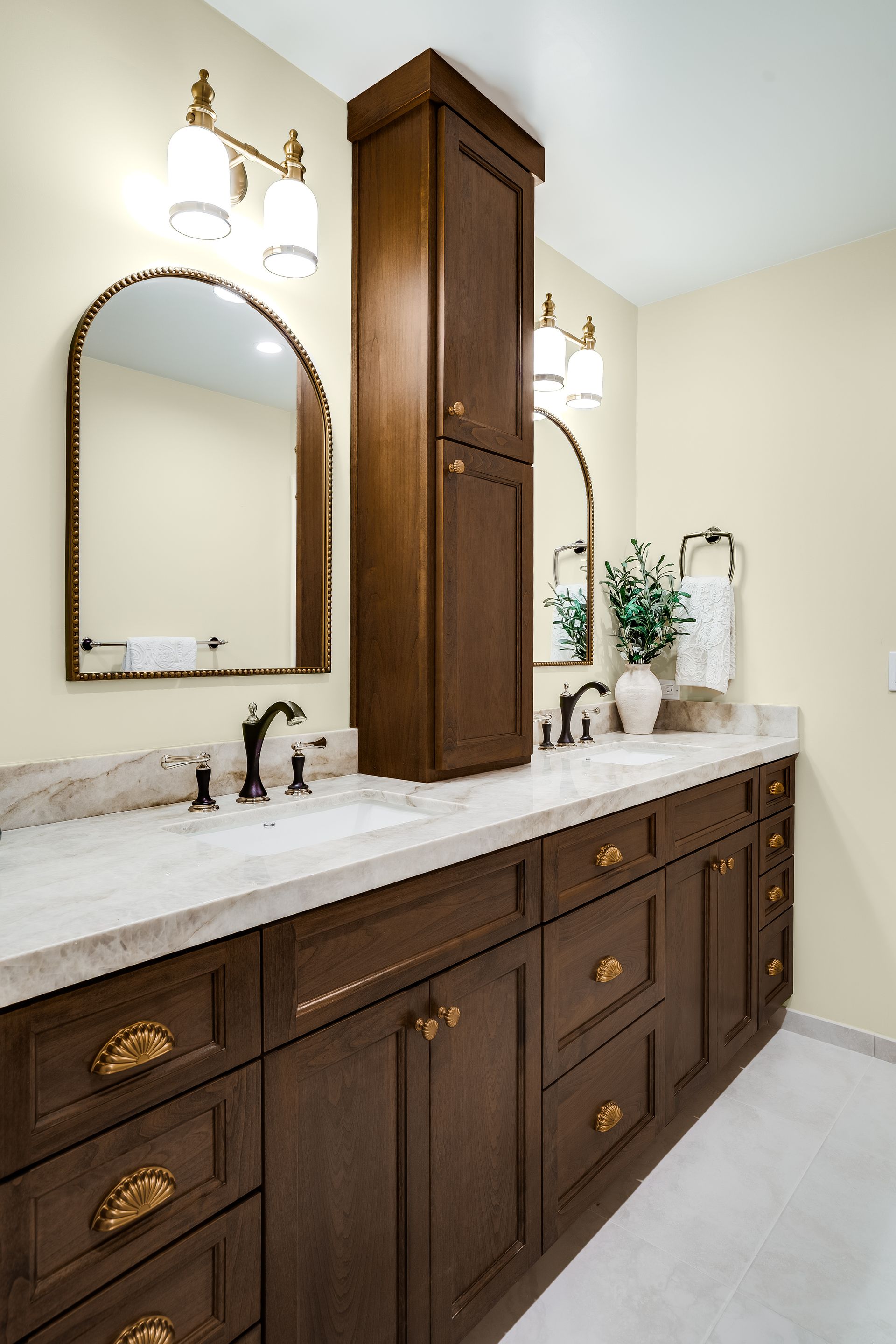 Bathroom with a long brown vanity, white countertop, arched mirrors, and tall cabinet.