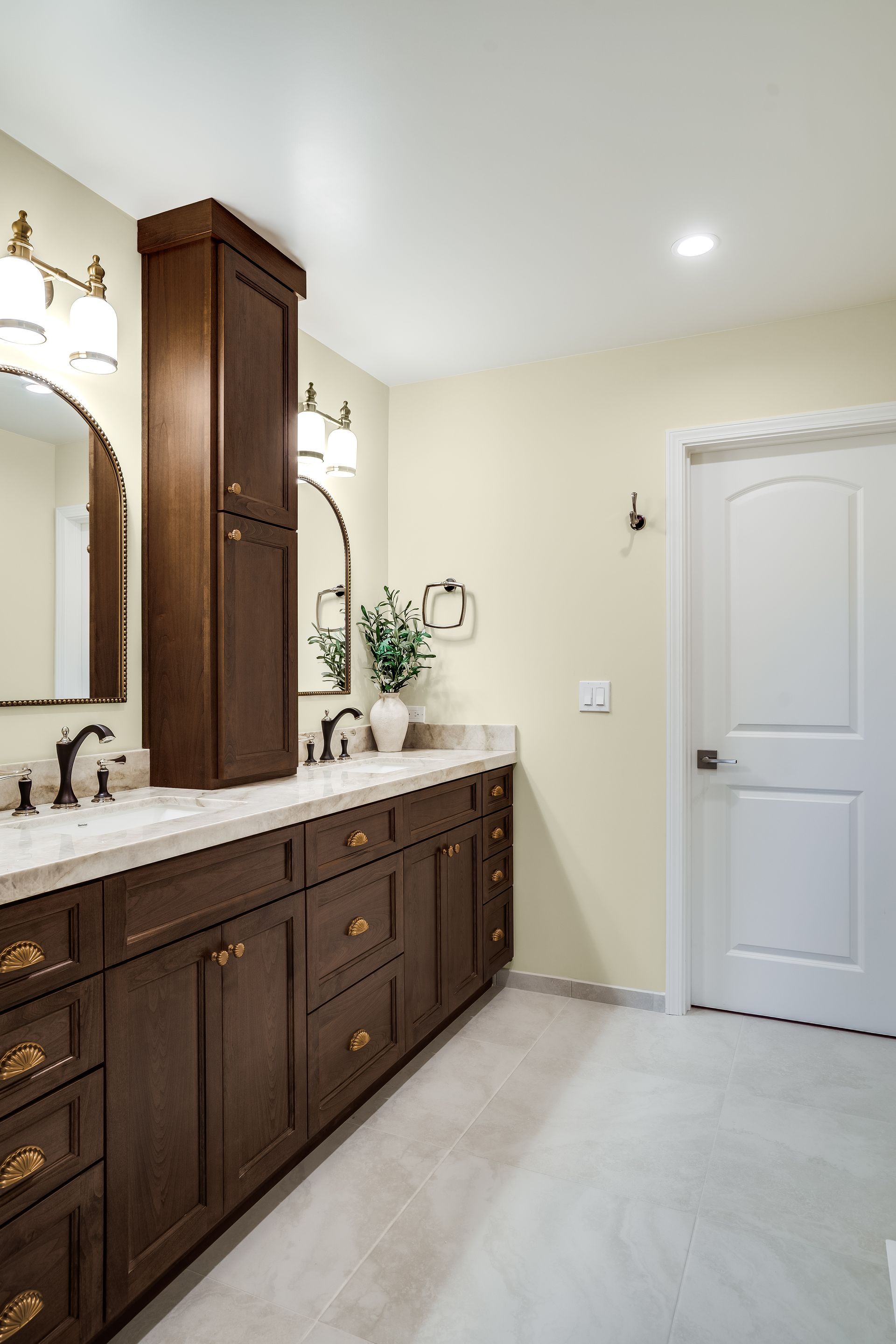 Dark brown vanity with gold hardware, light walls and floor, tall cabinet, mirrors, and a white door.