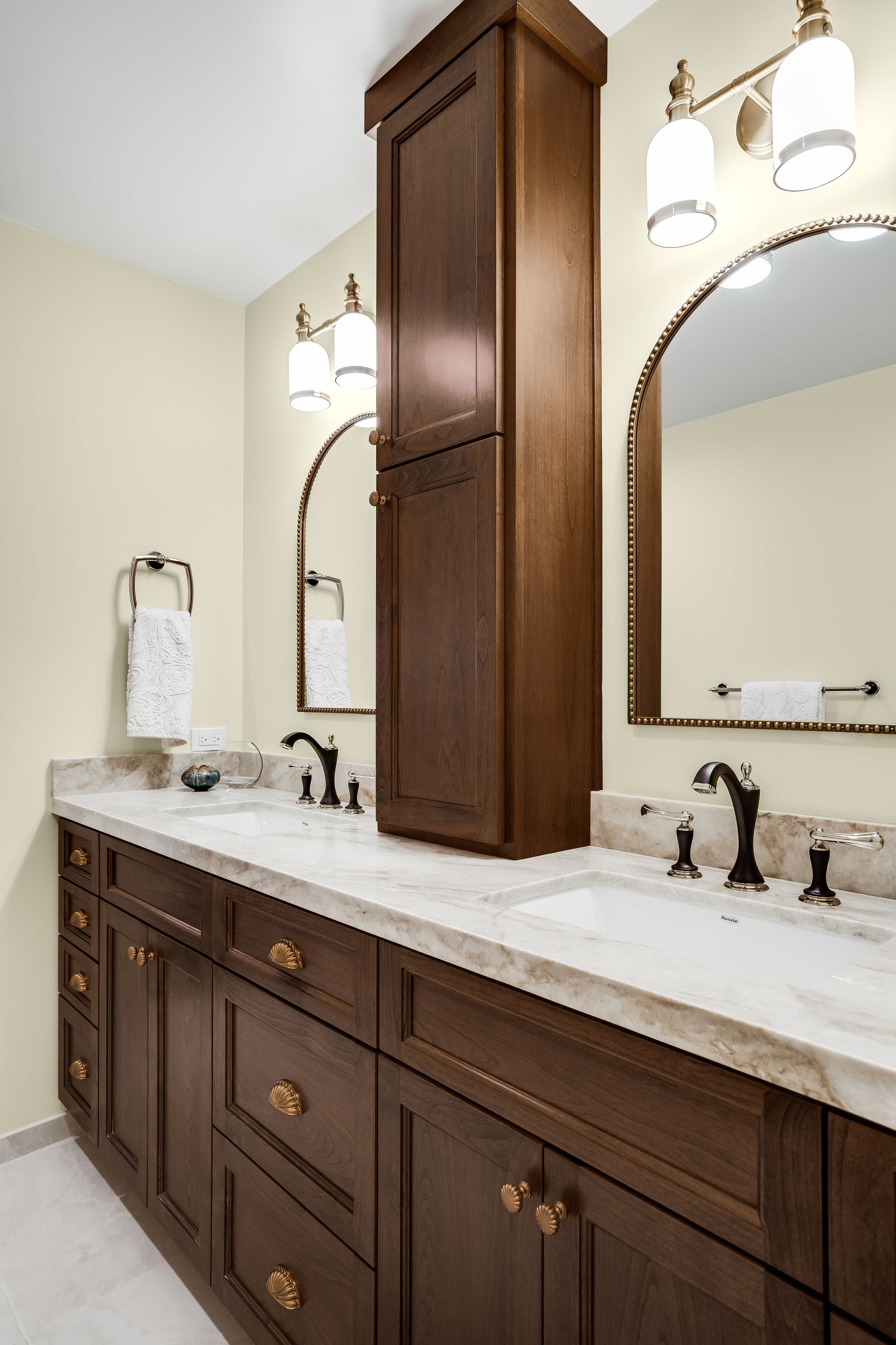 Bathroom with dark wood vanity, light countertops, and tall cabinet. Two arched mirrors and sconces.