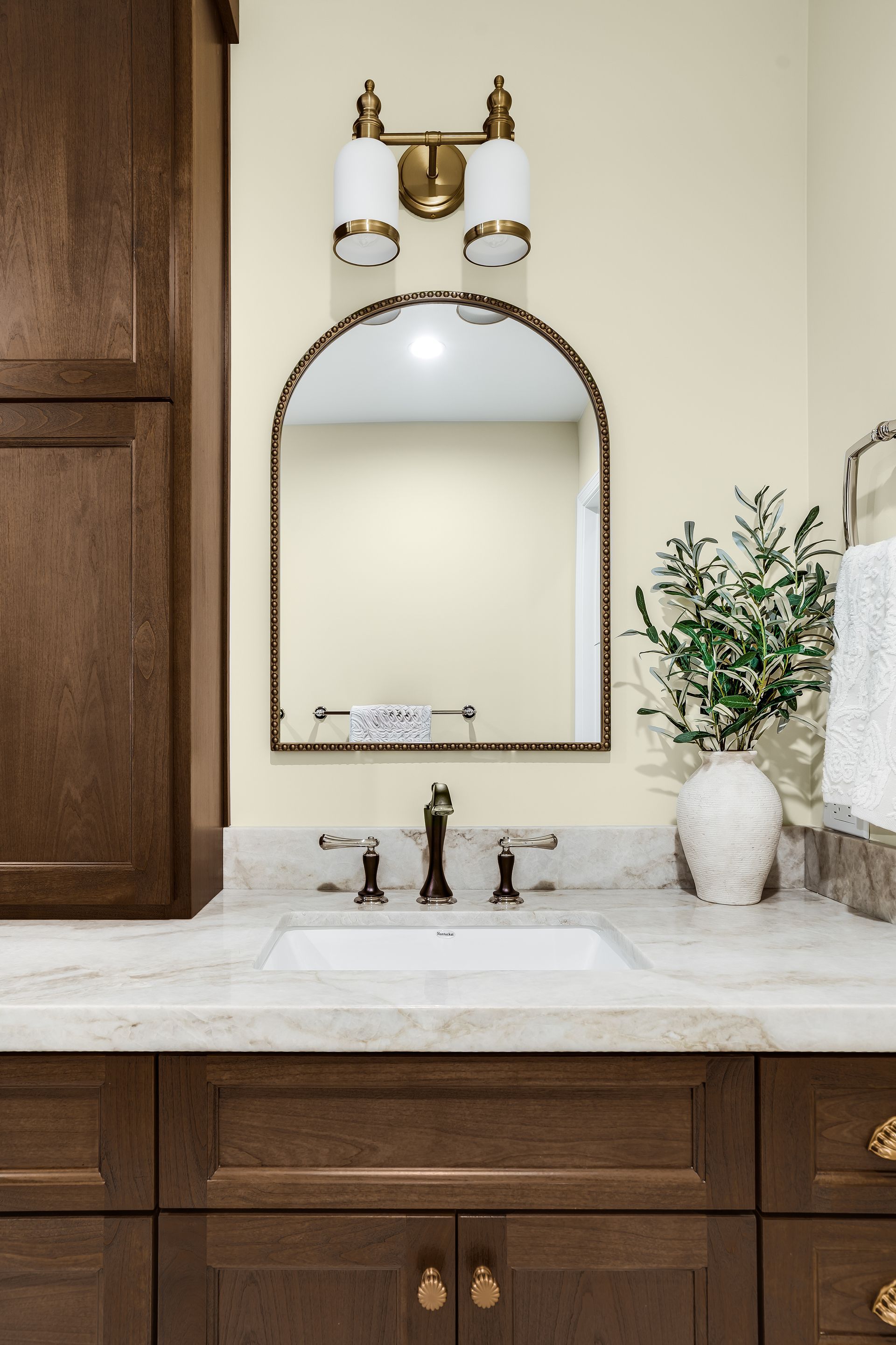 Bathroom vanity with brown cabinets, marble countertop, arched mirror, and brass fixtures.