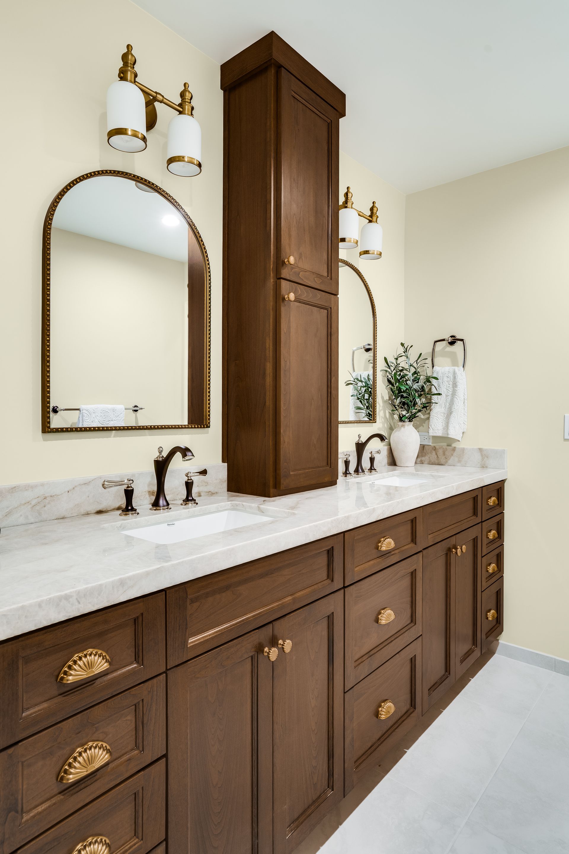 Bathroom with dark wood vanity, gold hardware, white countertop, arched mirrors, and cream walls.