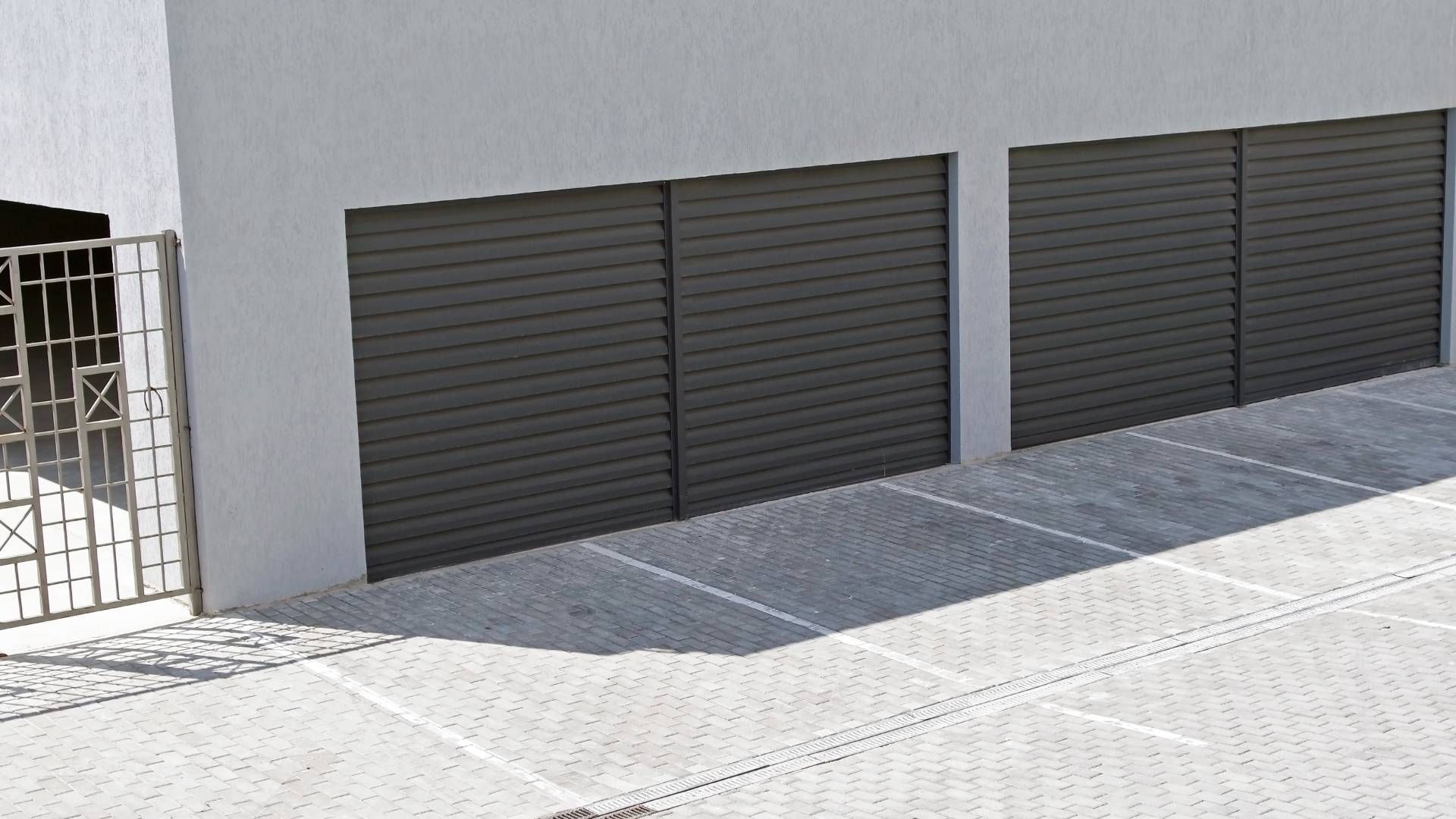 Two closed gray garage doors on a white building, with a gated entrance and a gray tiled driveway.