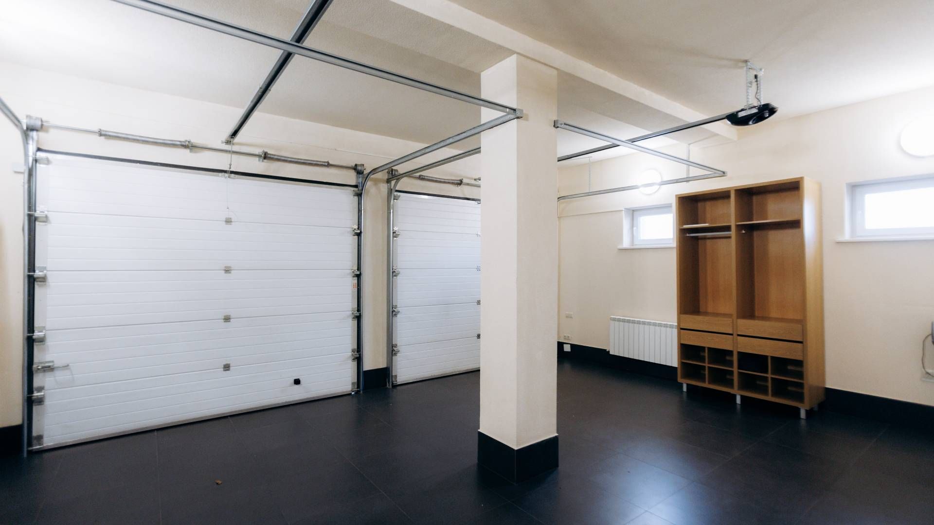 Two-car garage with tan doors, attached to a two-story beige house under a bright blue sky.