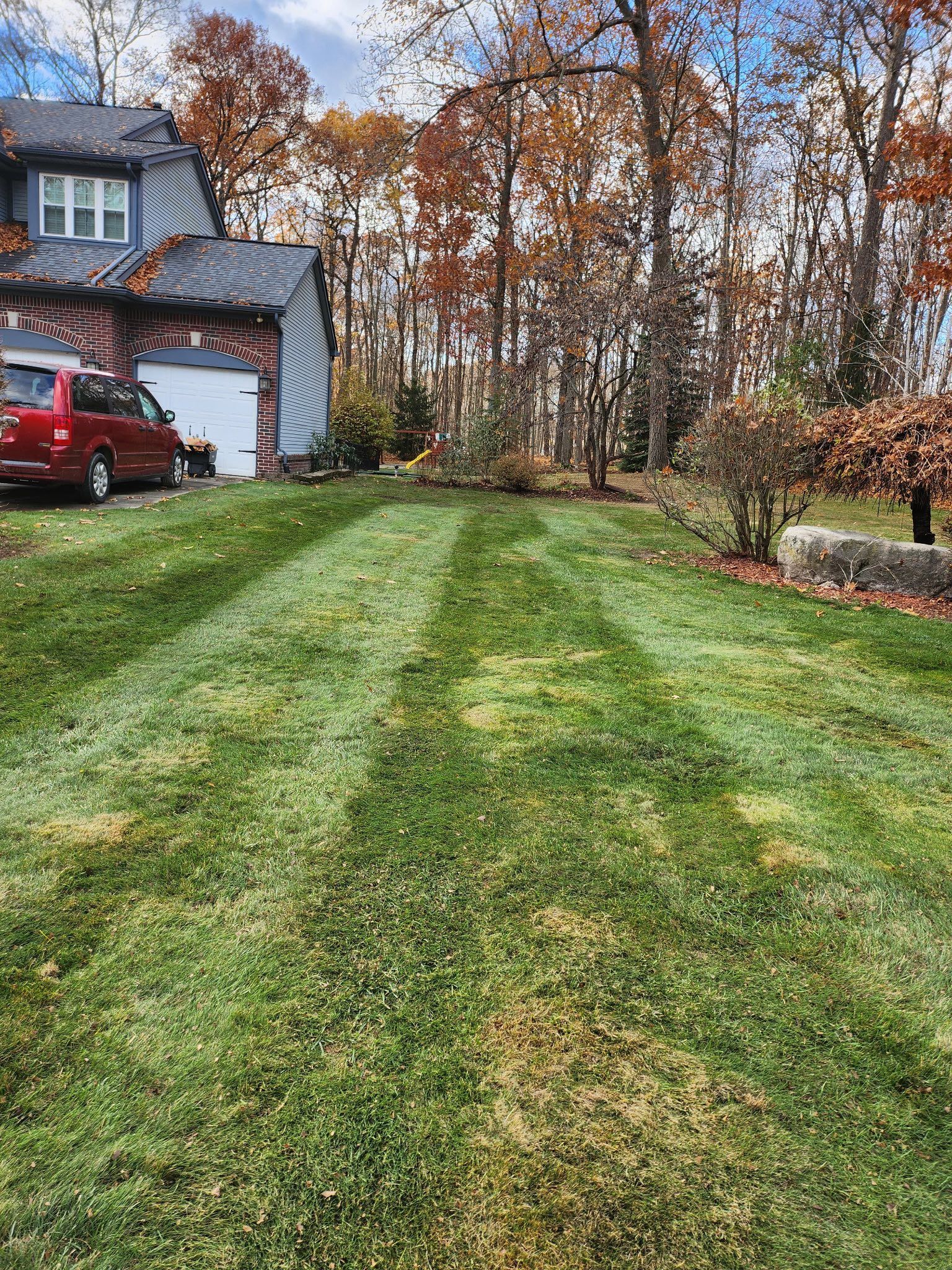Lawn with mowing stripes in front of a house, car in driveway. Trees with autumn leaves in the background.