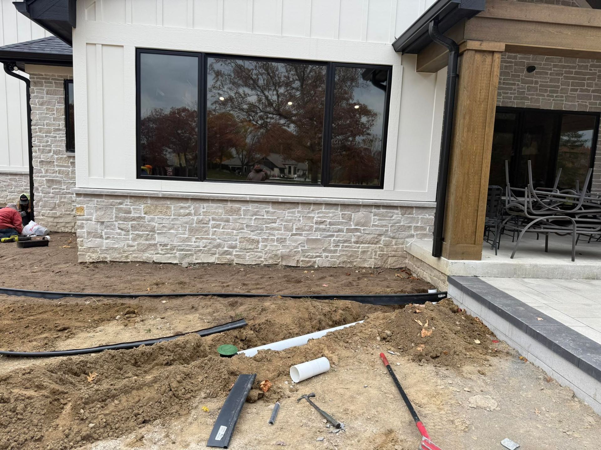 Exterior view of a house under construction with exposed dirt, tools, stone facade, and large windows.