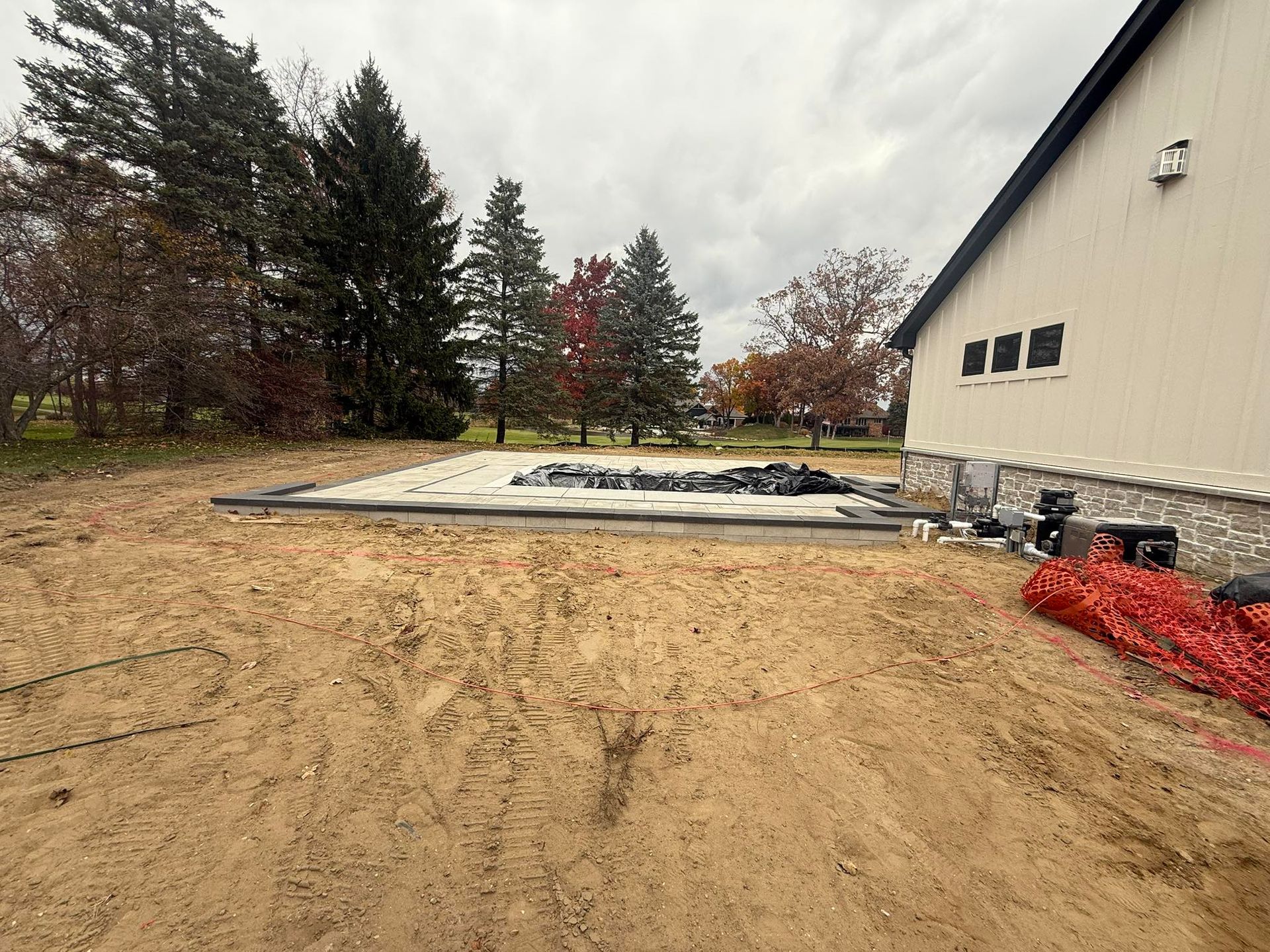 Construction site, beige building next to a sandy area; unfinished pool in background. Overcast sky.