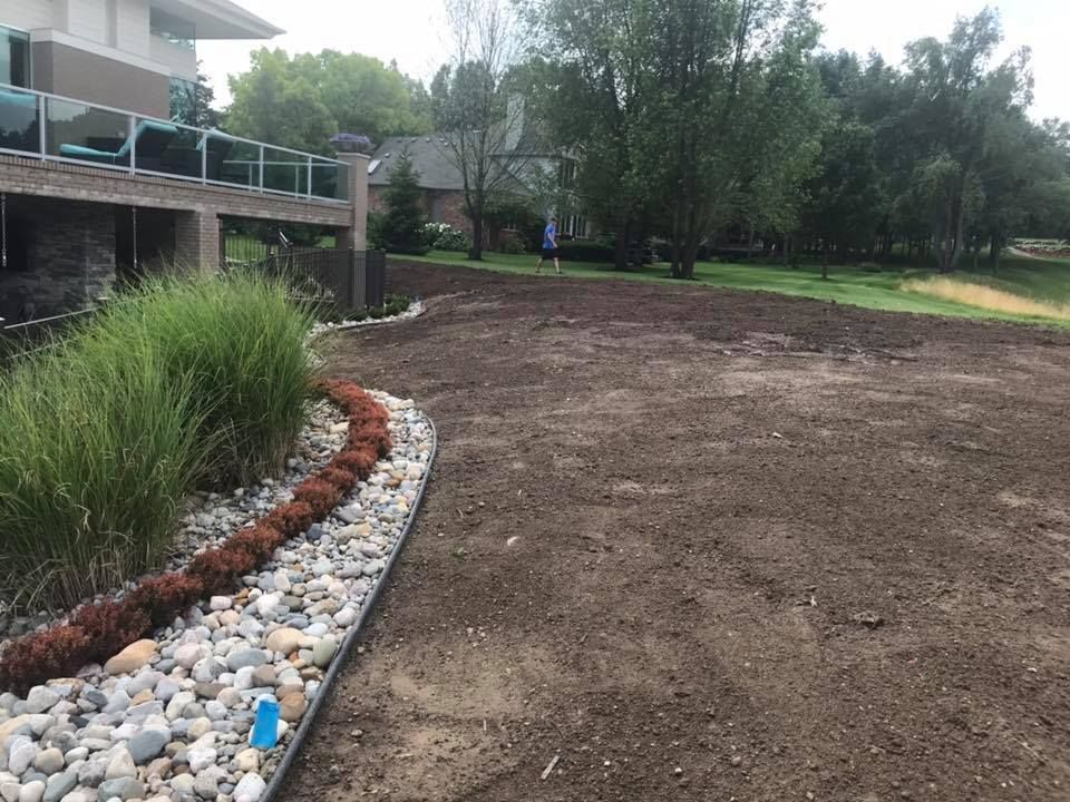 A backyard with freshly tilled soil, a landscaped border with rocks and plants, and a house with a deck.