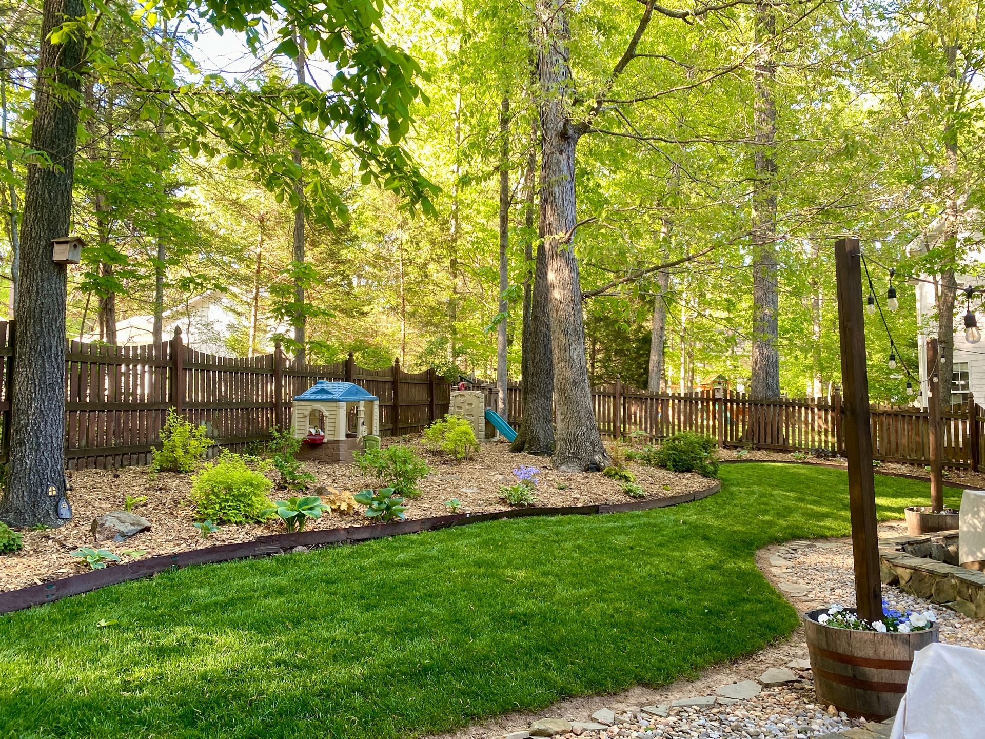 Lush green lawn borders a landscaped yard with a wooden fence, trees, and a small playhouse.