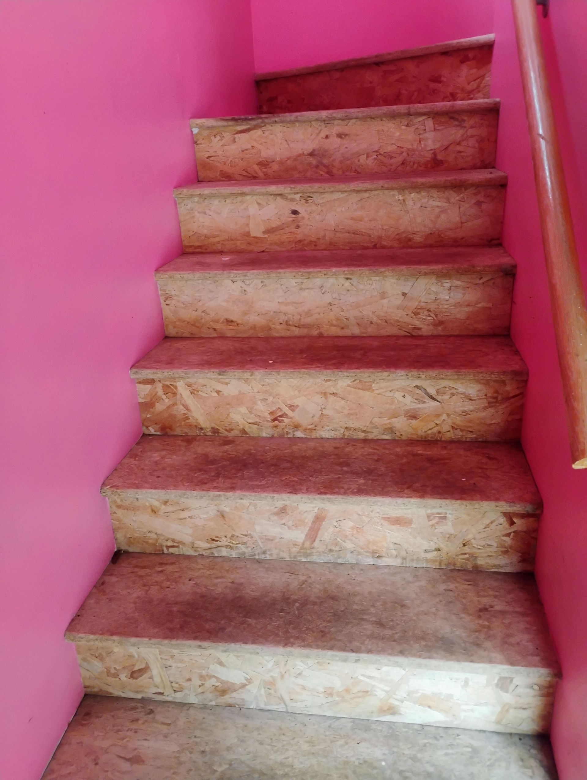 Staircase with light brown steps and pink walls, leading upwards. A wooden handrail is on the right.