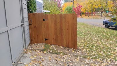 Wooden gate with black hinges, next to a light-colored building, with fall foliage visible.
