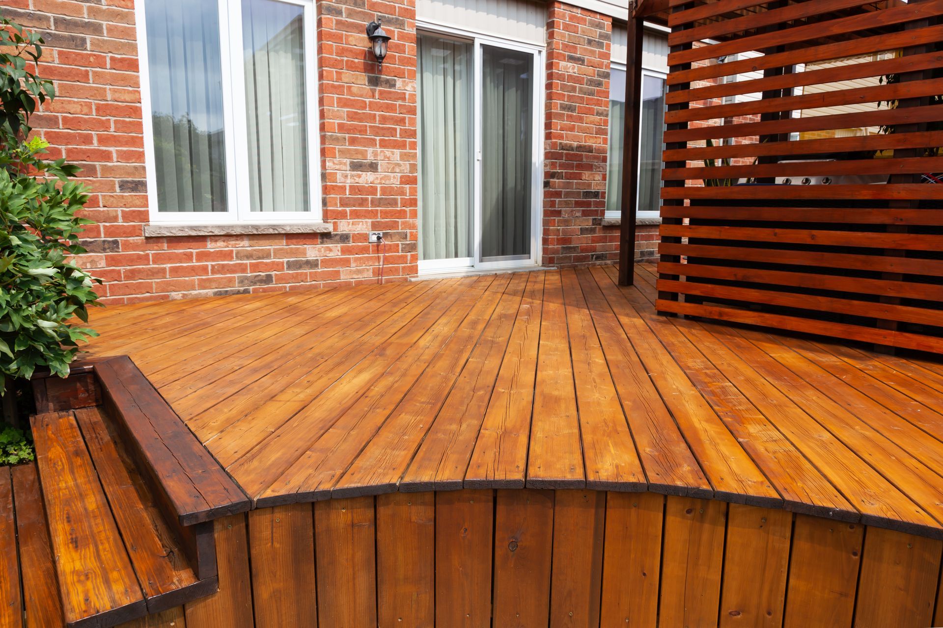 Wooden deck with curved edge, steps, and privacy screen against a brick building.