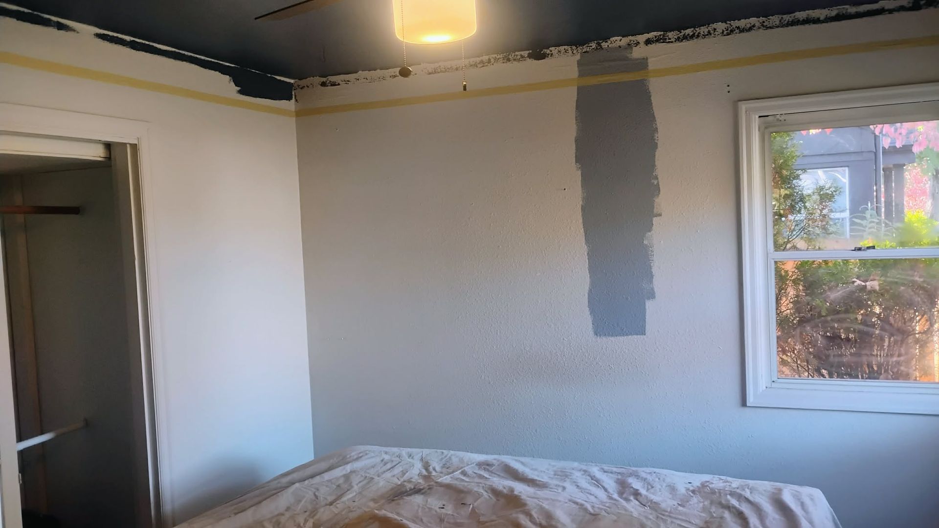 Bedroom interior: light-colored walls, gray paint swatch. Window on the right, open door on the left, unmade bed.