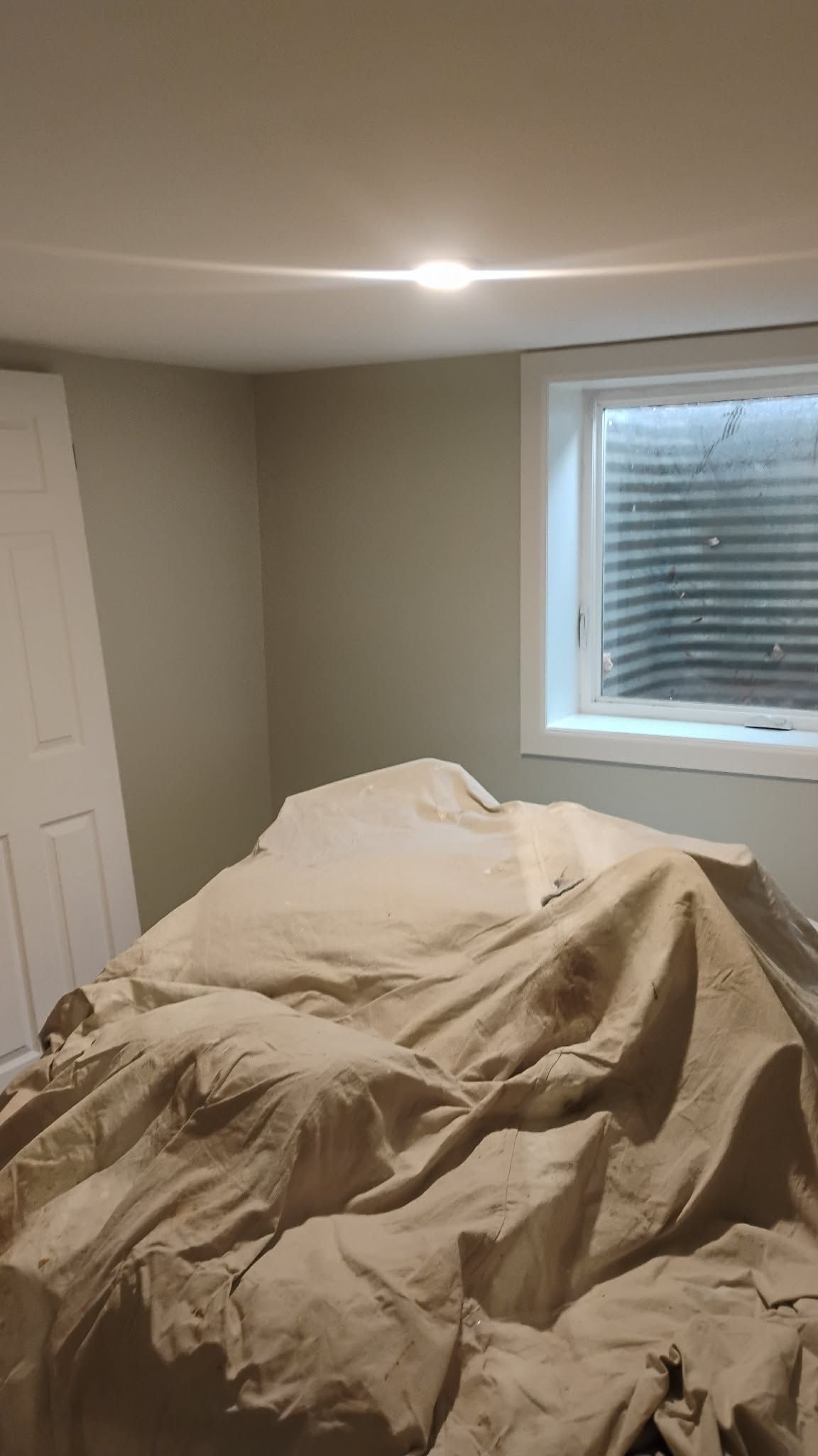 Bedroom interior with covered furniture, window, and door. Walls are painted light gray.