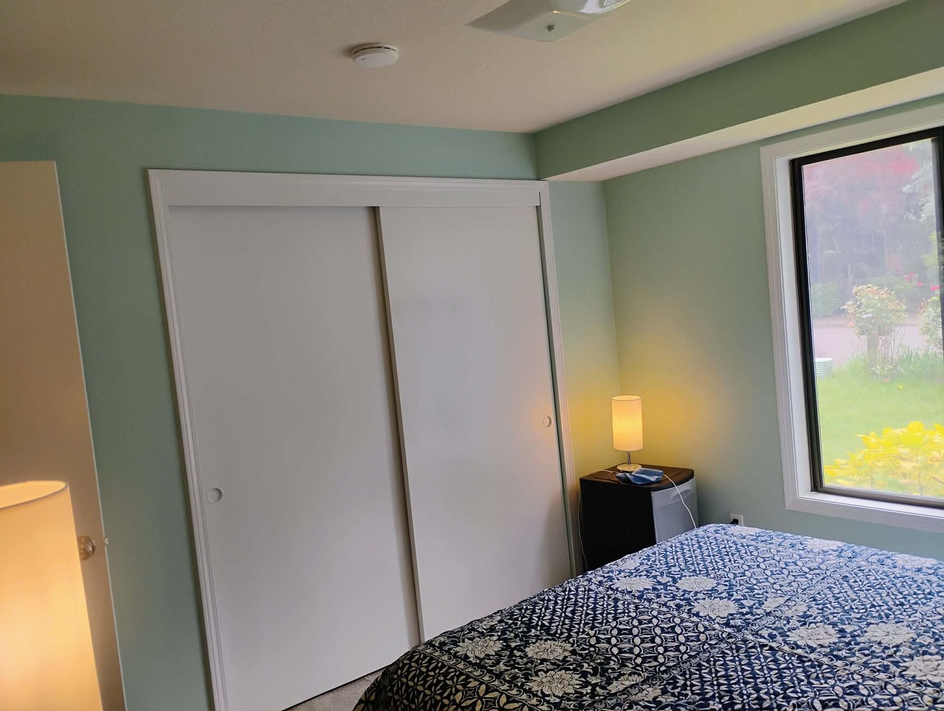 Bedroom with white closet doors, bed, nightstand, window, and light green walls.
