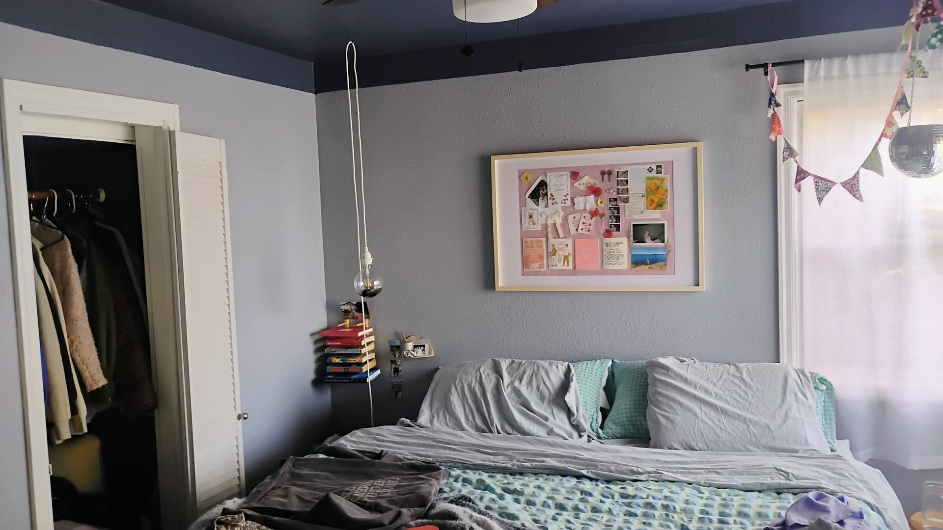 Bedroom with a bed, closet, framed art, string lights, and a window with a sheer curtain. Blue and gray tones.