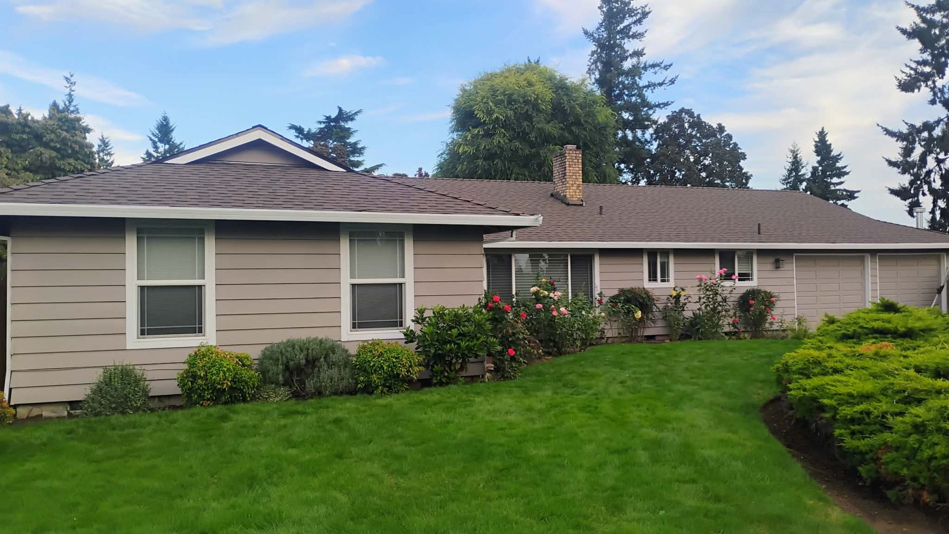 Tan house with brown roof and green lawn, bushes, and flowers in front. Cloudy sky.
