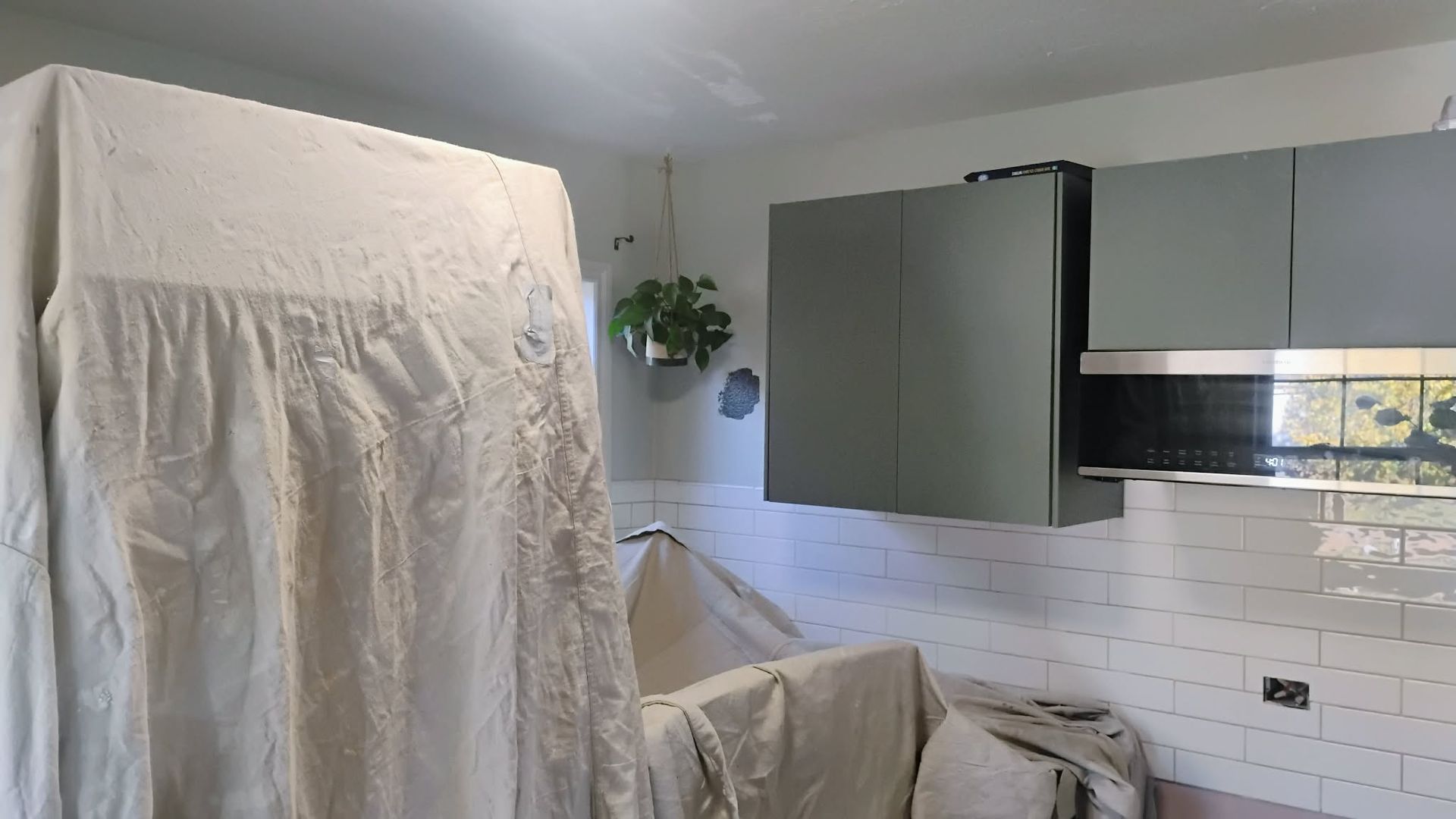 Kitchen renovation: cabinets (gray-green), covered appliances, white tile backsplash, hanging plant, and a window.