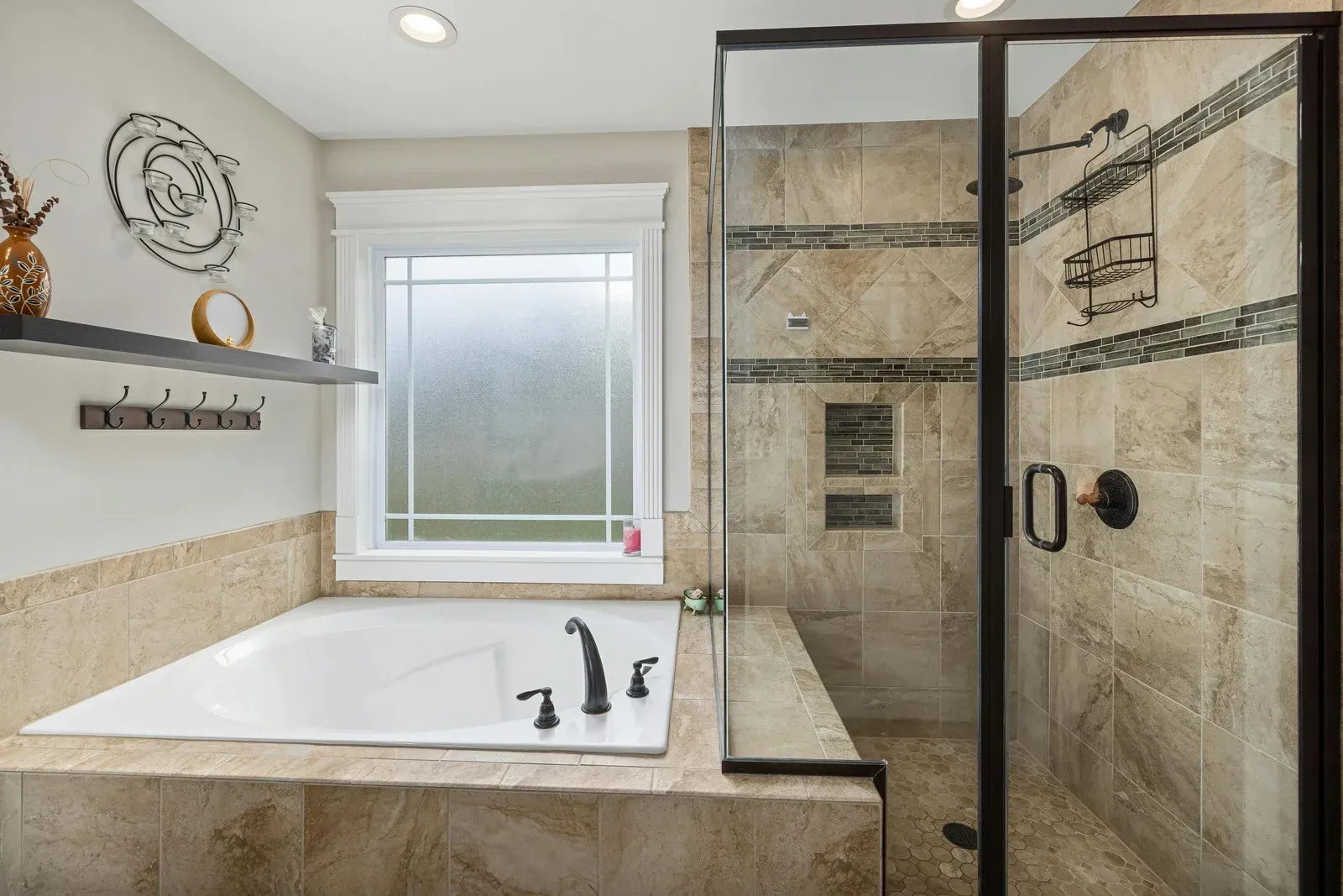 Bathroom with a bathtub, shower, and window. Beige tile, dark fixtures, and a decorative shelf are visible.