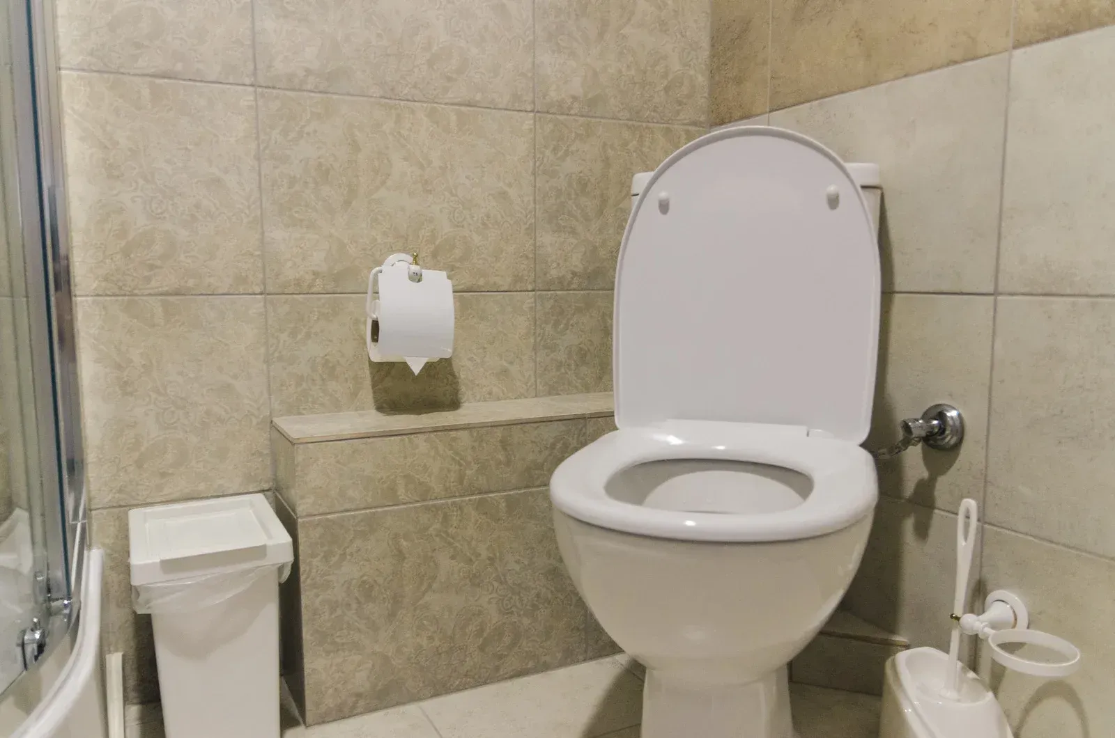Toilet in a bathroom with a trash can, toilet paper, and brush against beige tile walls.