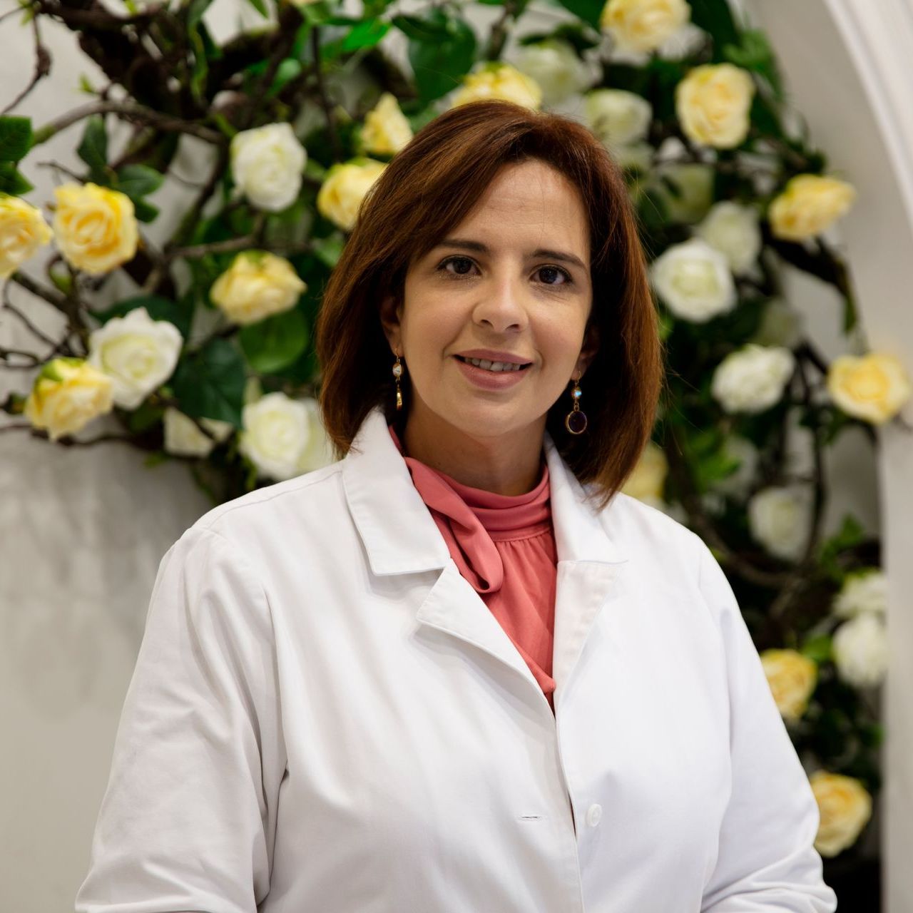 A woman in a white lab coat is standing in front of a wall of flowers.