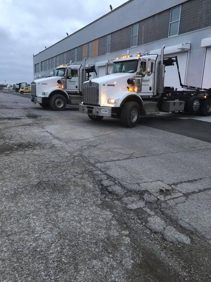 Two white dump trucks parked on cracked pavement in front of a gray industrial building.