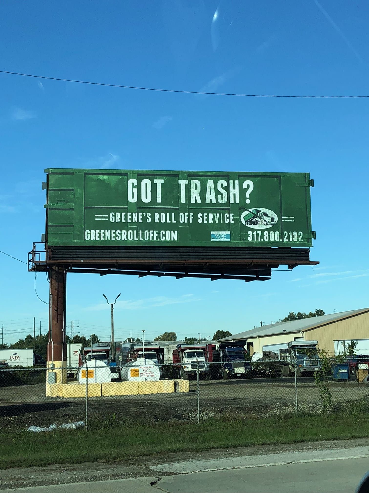 Billboard, 'GOT TRASH?' Green container, 'Greene's Roll Off Service'. Buildings, vehicles, clear sky.