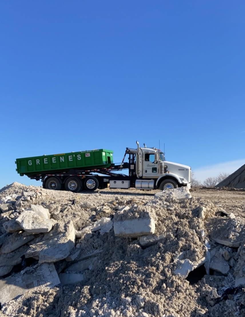 Green dump truck with green container on a debris pile under a blue sky.