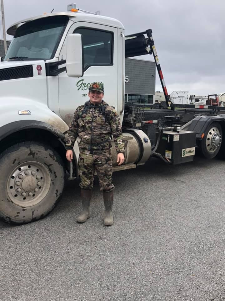 Person in camouflage standing next to a white industrial truck. Gray gravel lot.