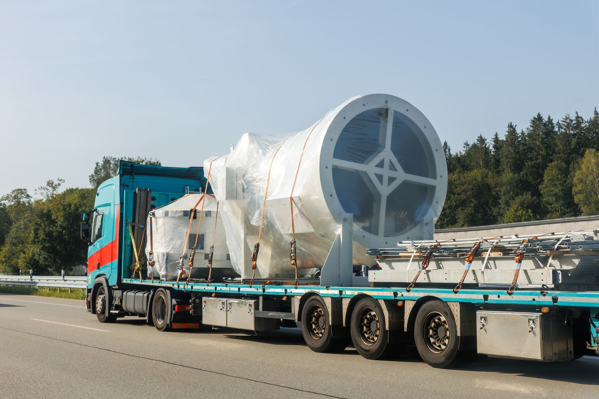 A large white industrial machine, wrapped and secured on a blue flatbed truck on a road, trees in background.