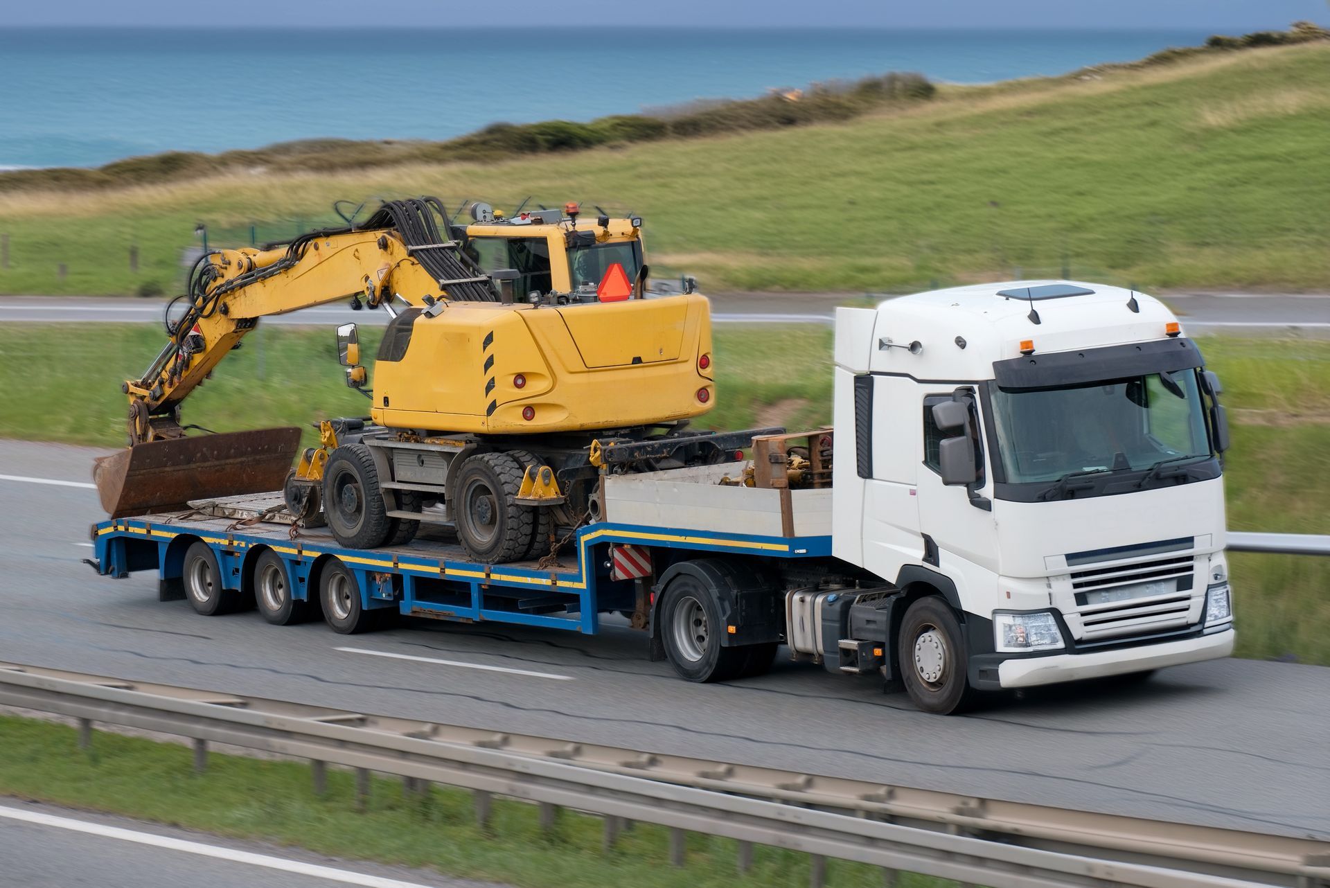 Yellow excavator on a flatbed truck traveling on a highway, with a green hill and ocean in the background.