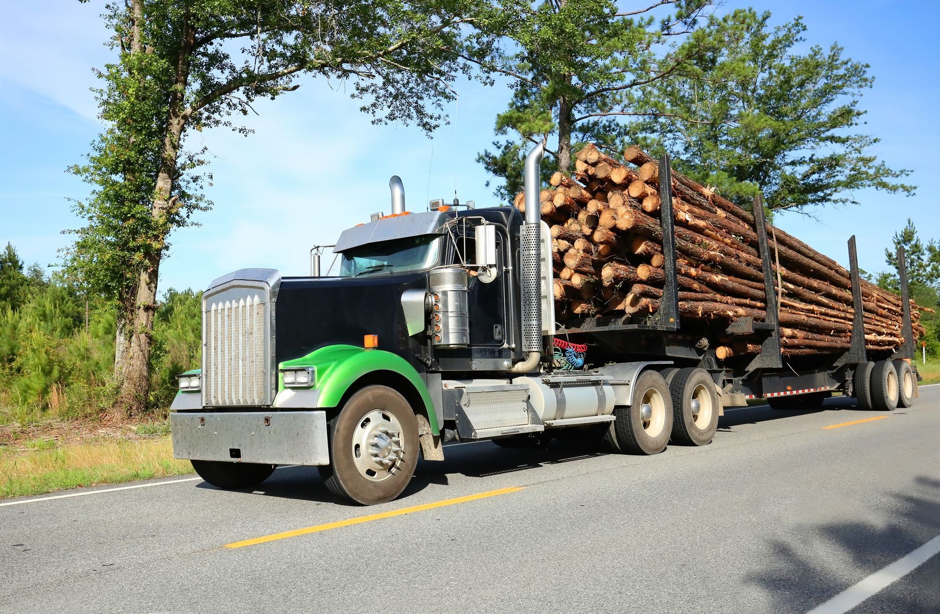 Black semi-truck hauling a load of logs on a paved road with trees on either side.