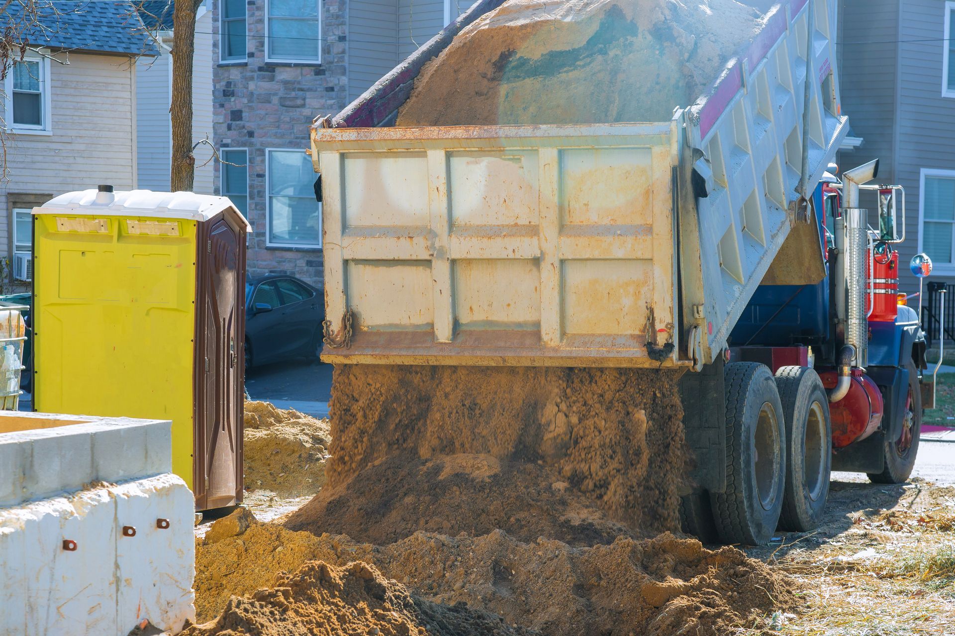Dump truck unloading dirt near a yellow portable toilet and a concrete structure.