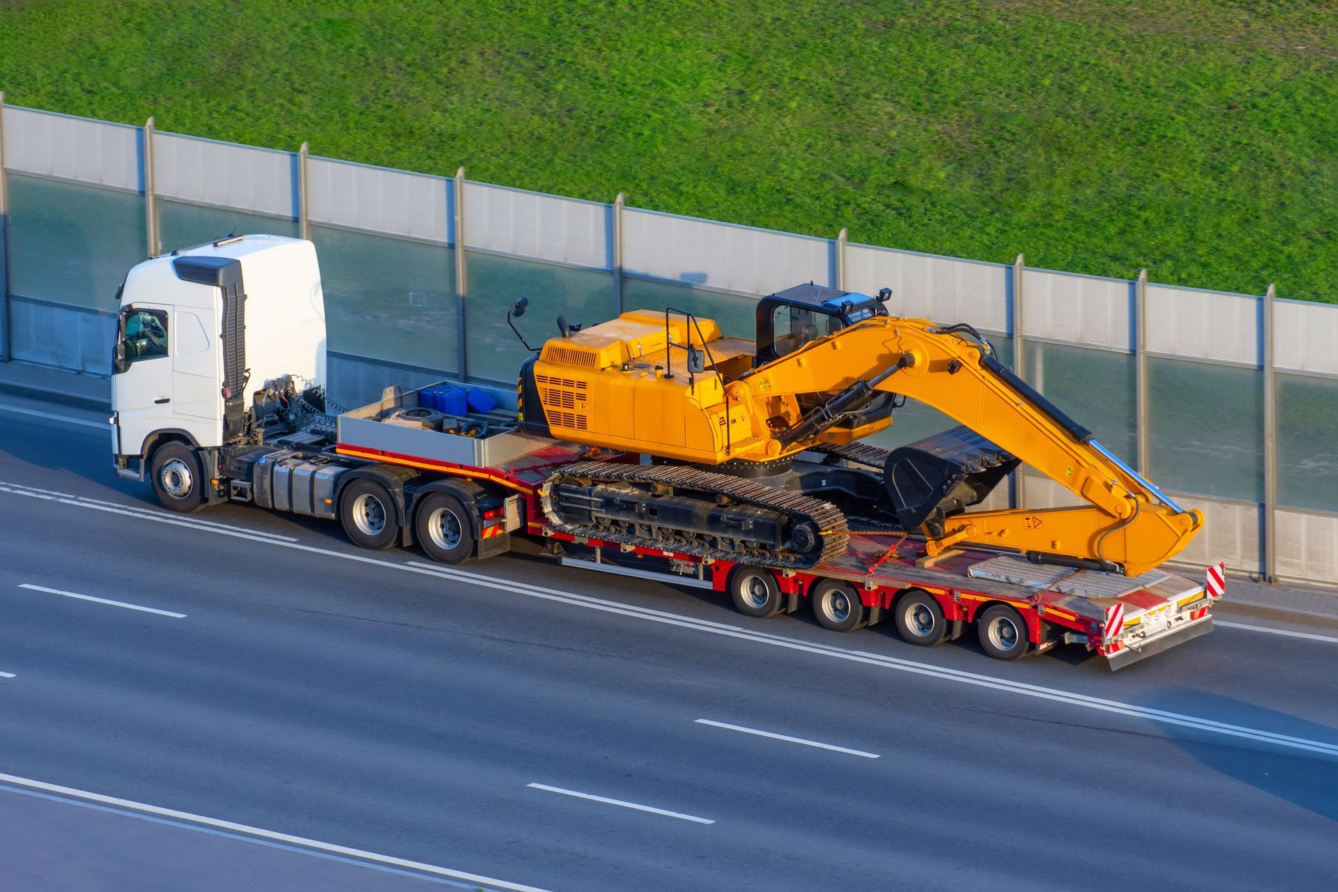 Yellow excavator on a flatbed truck traveling on a highway, next to a sound barrier.